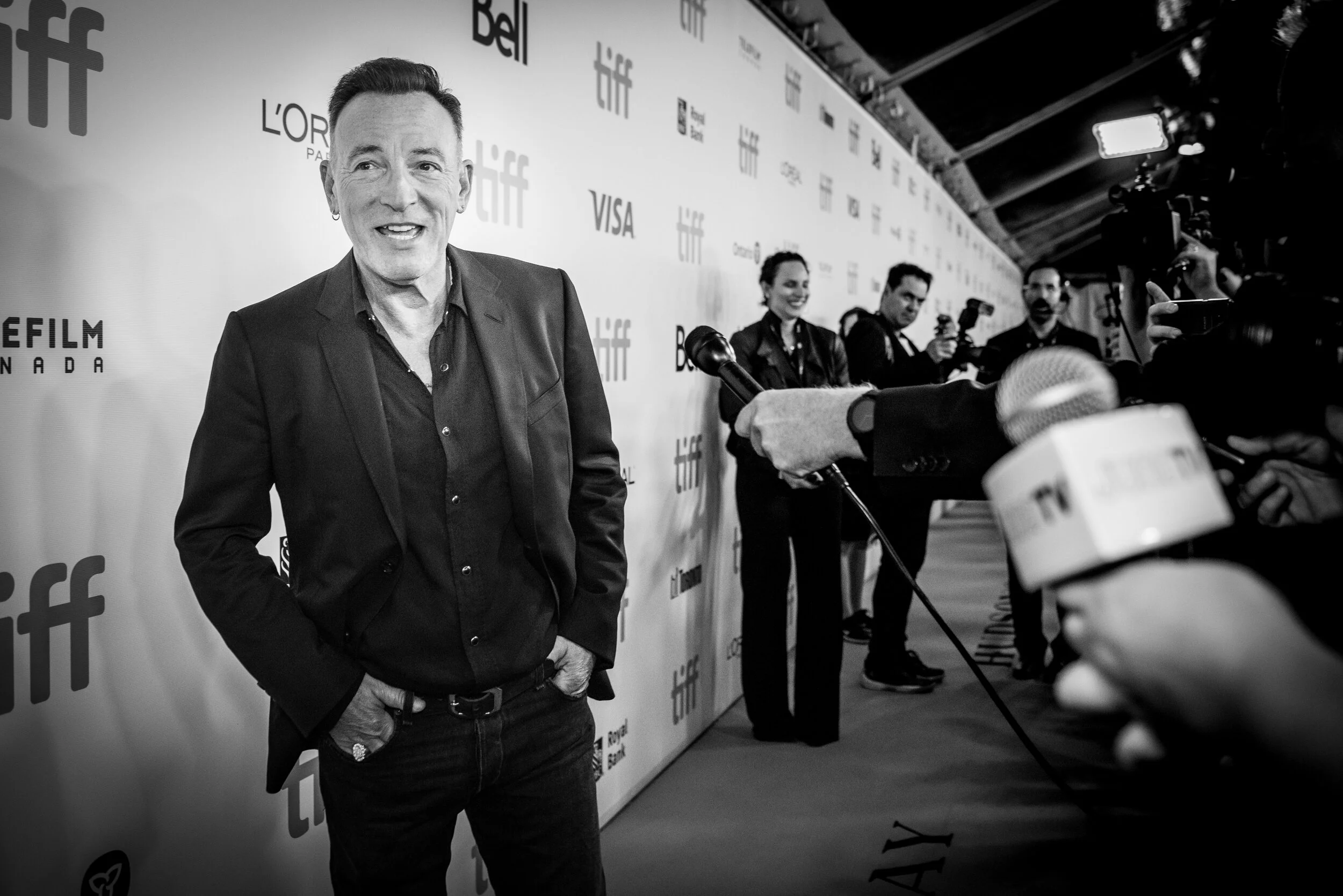 A man in a dark blazer and shirt smiling for the camera at a red carpet event, with a backdrop featuring logos of TIFF and other sponsors. Several people and photographers are in the background.