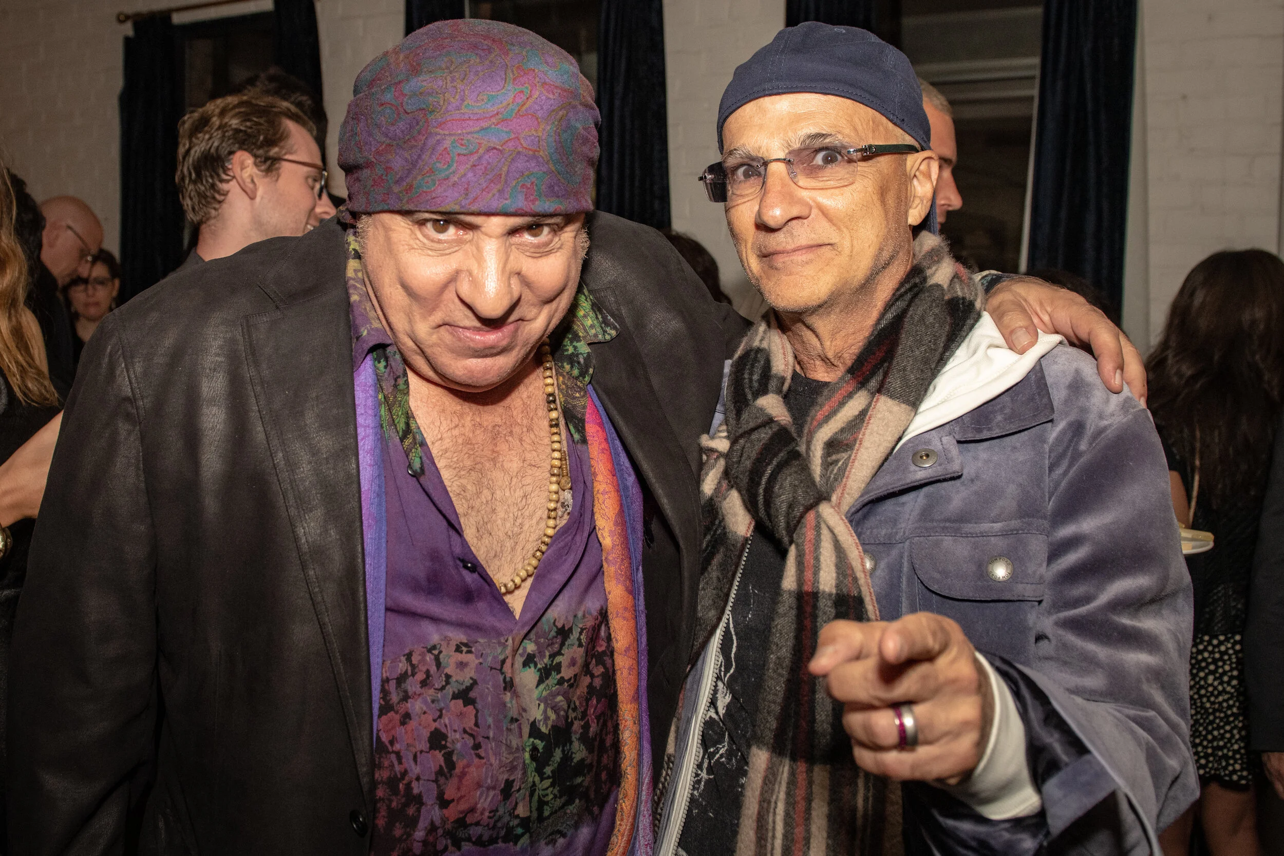 Two men pose together at a social gathering. The man on the left wears a purple shirt, a leather jacket, and a colorful headscarf; the man on the right wears glasses, a blue beanie, a plaid scarf, and a jacket. They are standing close, with the man o