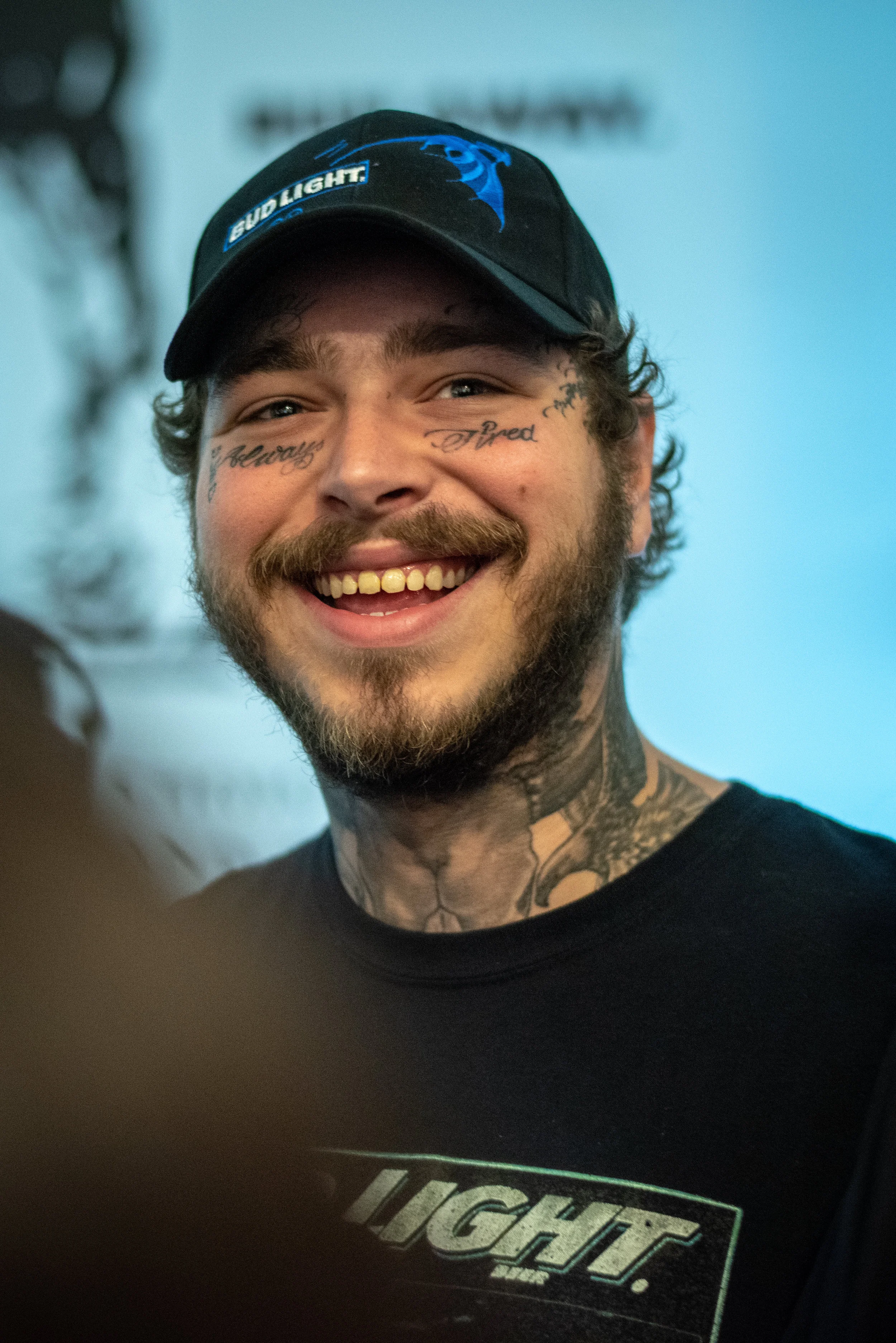 A smiling young man with a beard, tattoos on his face and neck, wearing a black baseball cap and a black t-shirt.