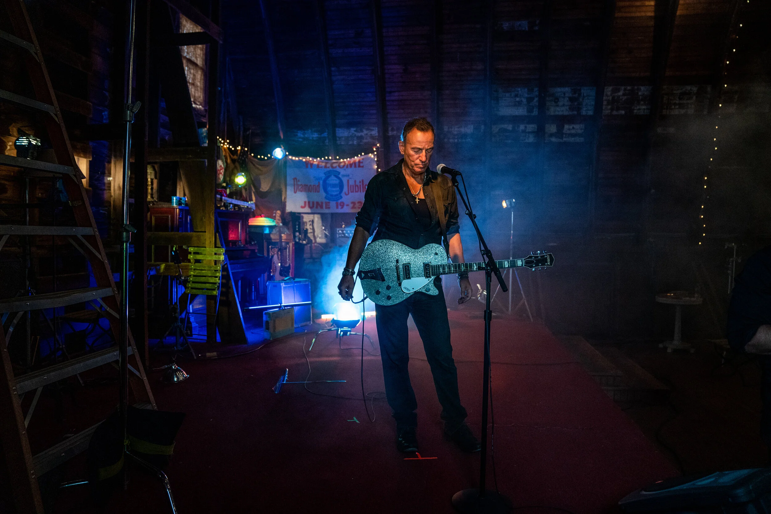 A man holding a guitar on stage with colorful lighting in a rustic indoor venue, a banner in the background reading 'Welcome Diamond Jubilee June 19-23'.