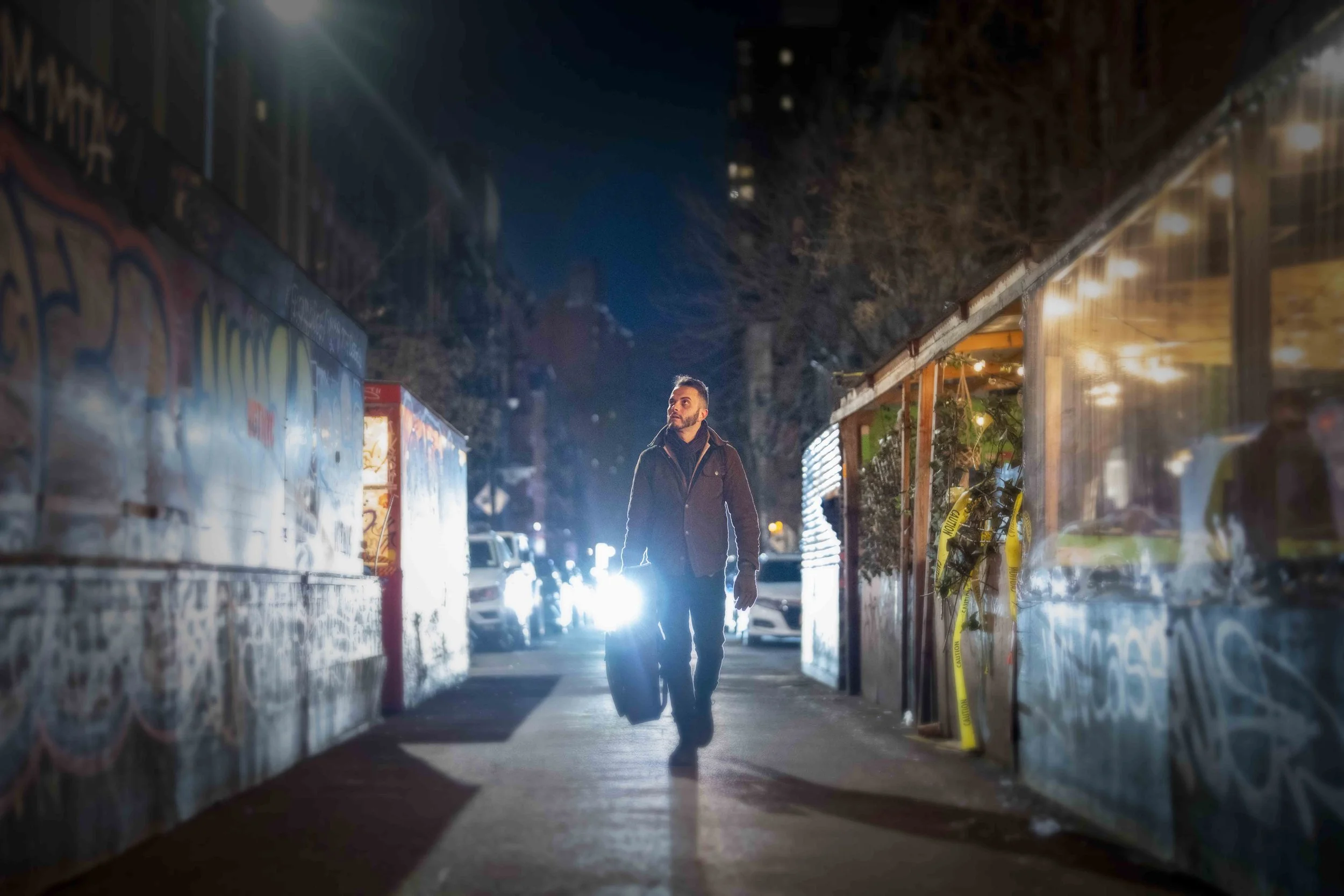 A man walking on a city sidewalk at night, carrying a small suitcase, with cars and bright lights behind him.
