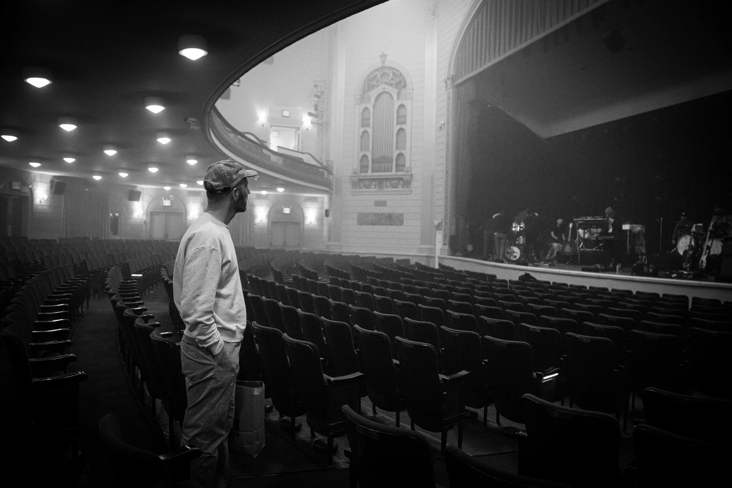 A man wearing a cap and casual clothing stands alone in an empty theater auditorium, looking toward the stage. The theater has rows of chairs, ornate interior details, and a large stage with musical instruments and equipment set up, ready for a perfo