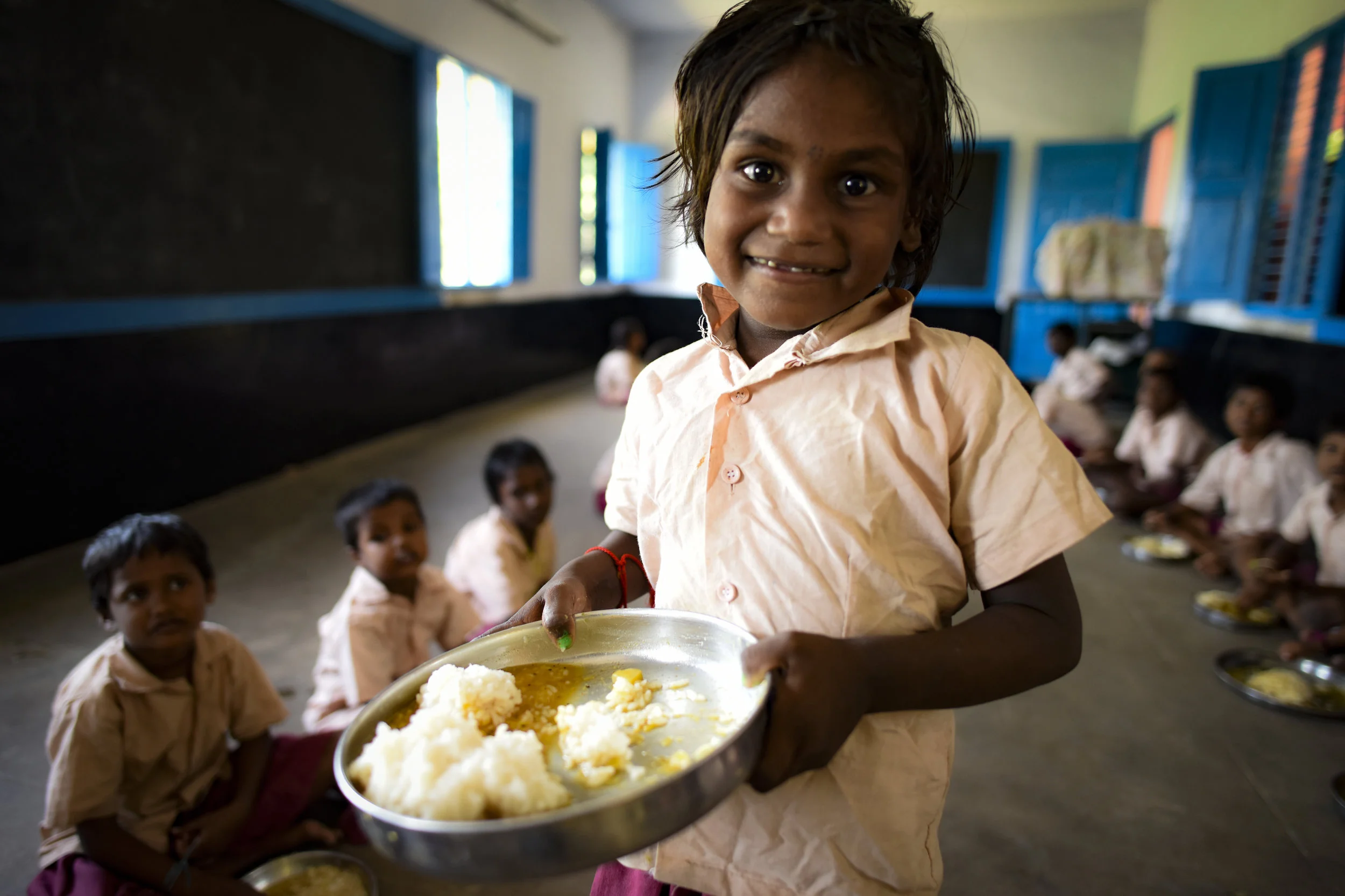 A young girl in a school uniform holding a tray of food, standing in a classroom with other children sitting on the floor behind her.