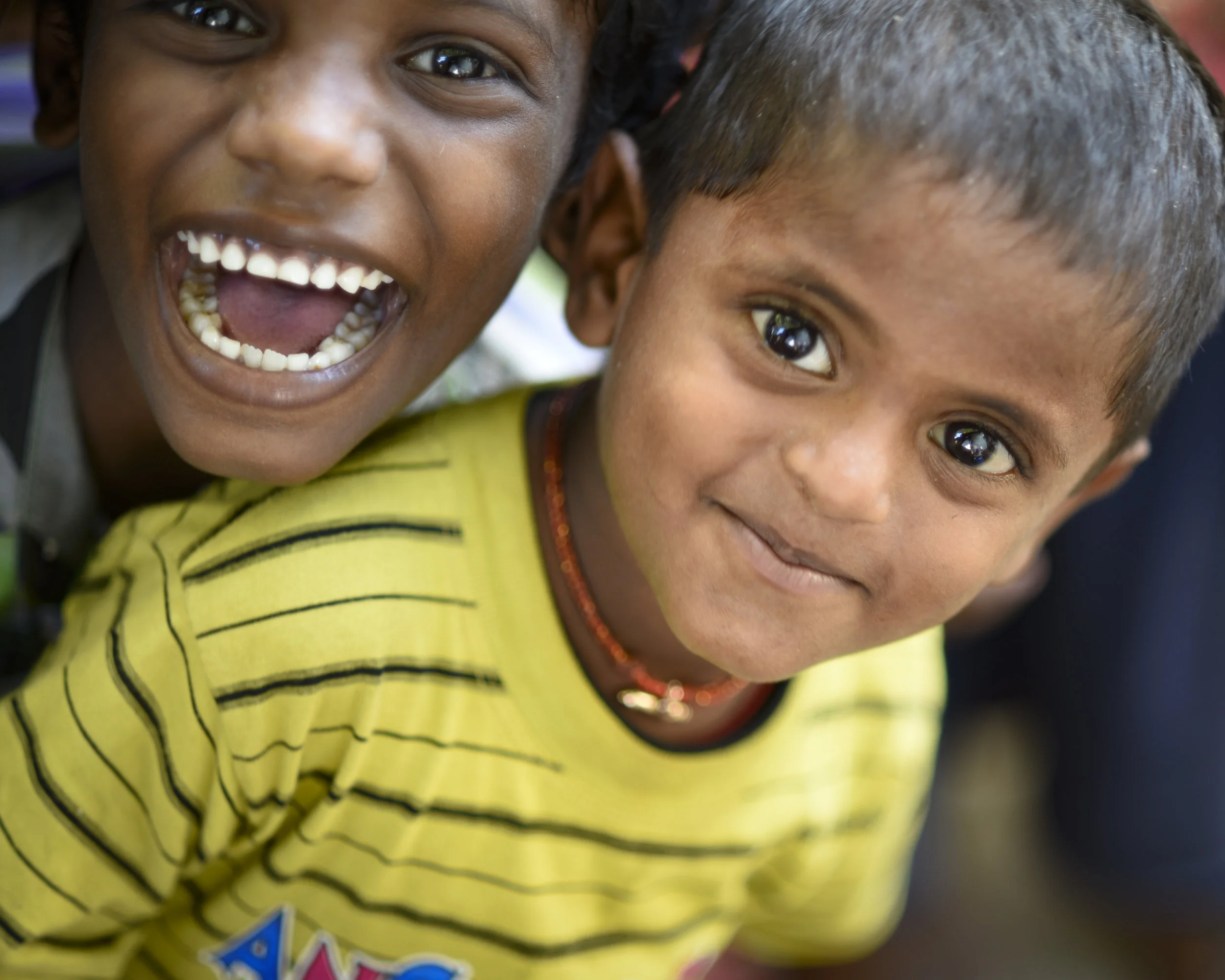 Two young children with dark hair and brown skin, one with a big open-mouthed smile showing teeth, the other with a subtle smile, both wearing yellow shirts with black stripes, close together in a cheerful moment.
