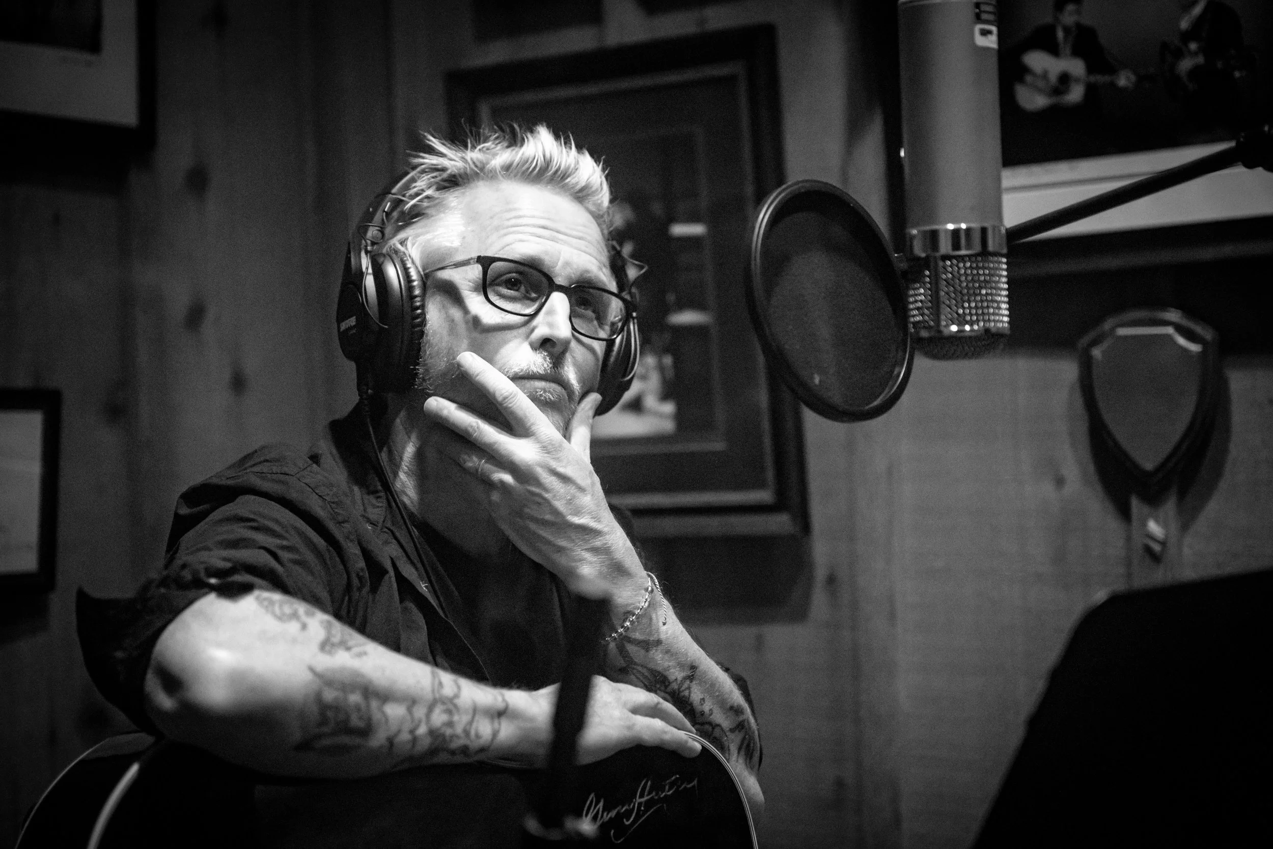 A man with glasses, tattoos, and a beard, wearing headphones, sitting in front of a microphone in a recording studio, holding his chin with one hand while looking thoughtfully at a music stand or screen.