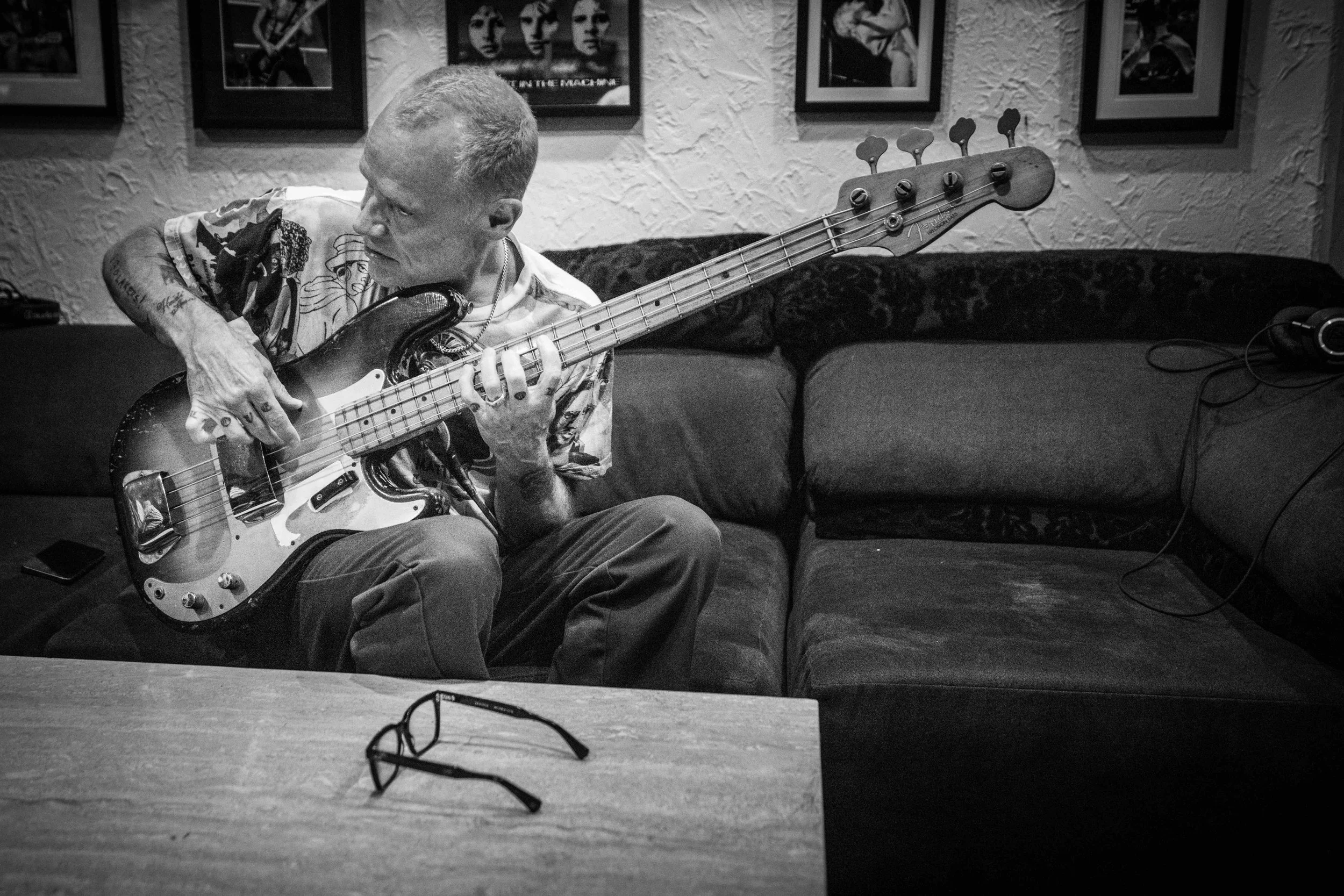 A man sitting on a couch, playing an electric bass guitar. He has tattoos, short hair, and is wearing a patterned short-sleeve shirt. On the table in front of him, there is a pair of eyeglasses. The background features posters on the wall.