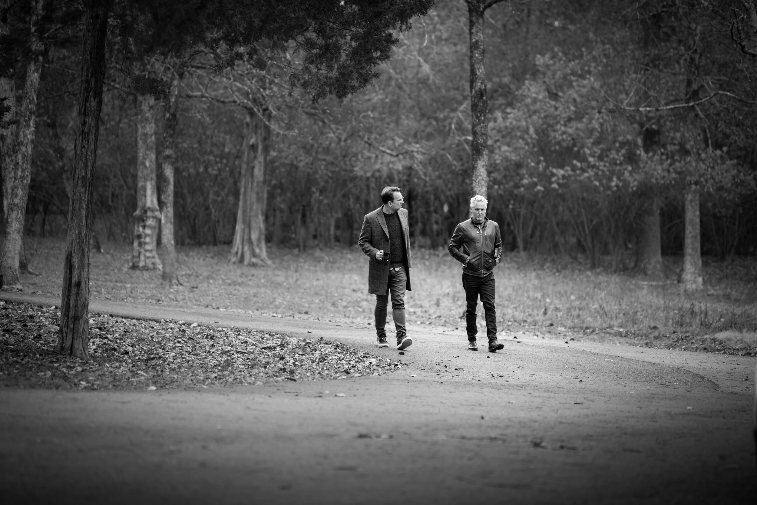 Two people walking on a path through a wooded park, one holding a smartphone, in black and white.