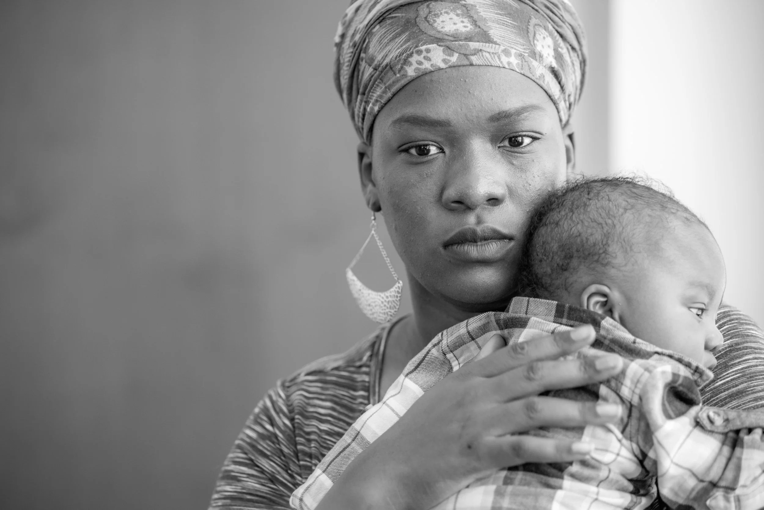 A woman with a patterned headwrap and earring holding a baby, looking directly at the camera in black and white.