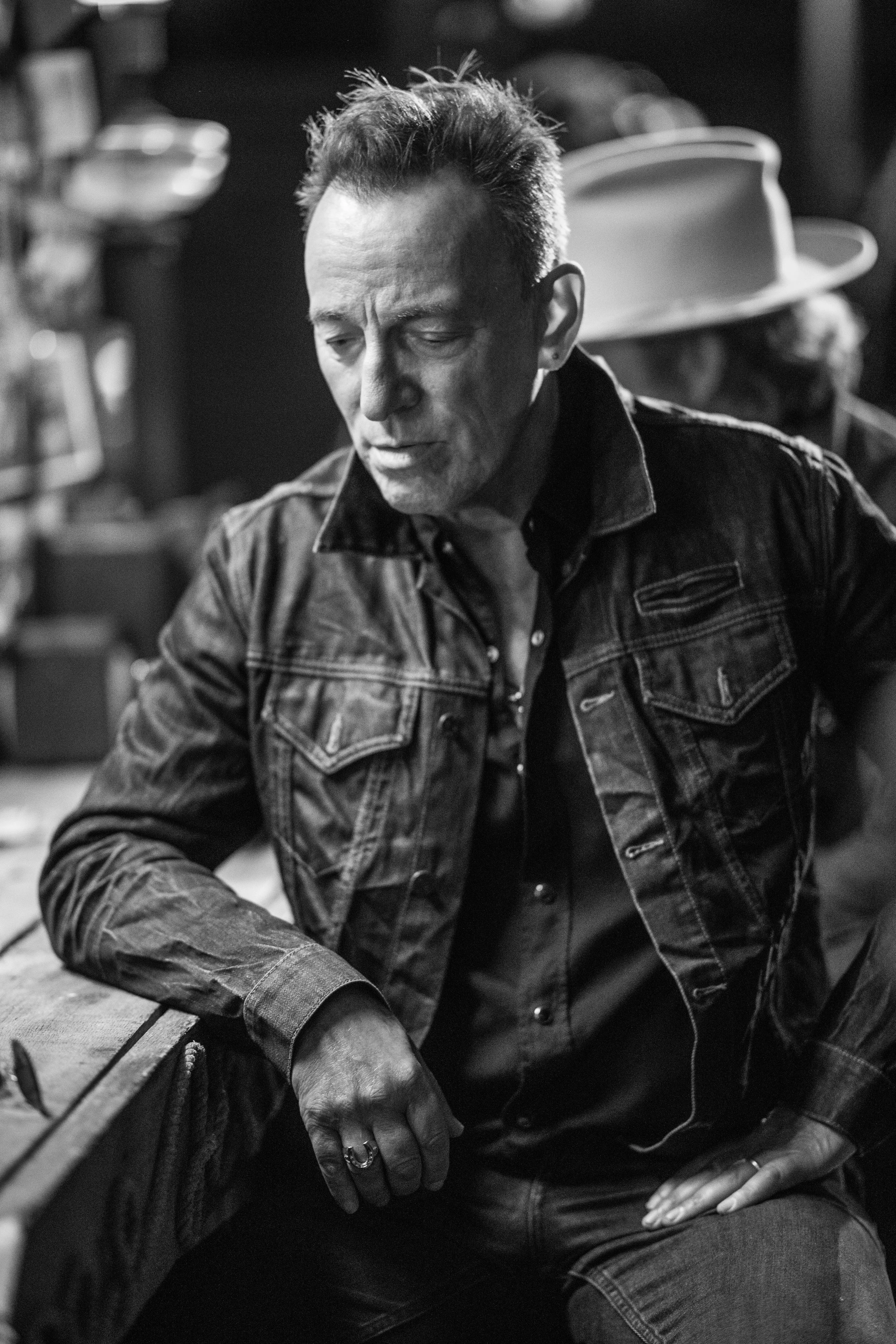 Black and white photo of a man with styled hair, wearing a denim jacket and a ring, sitting at a table with a person in a hat in the background.