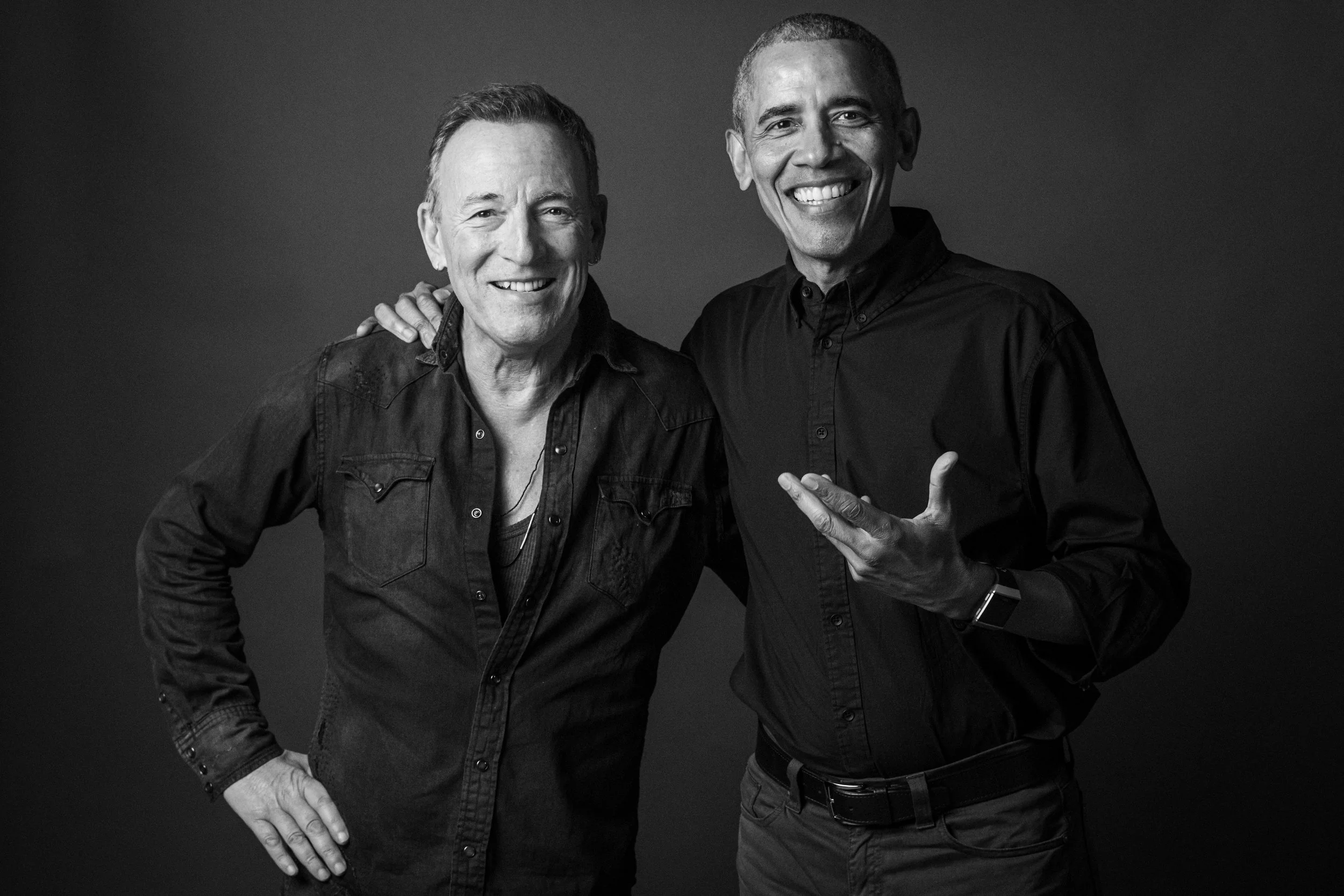 Two smiling men standing close together, one with his hand on his hip and the other pointing at him, against a plain dark background.