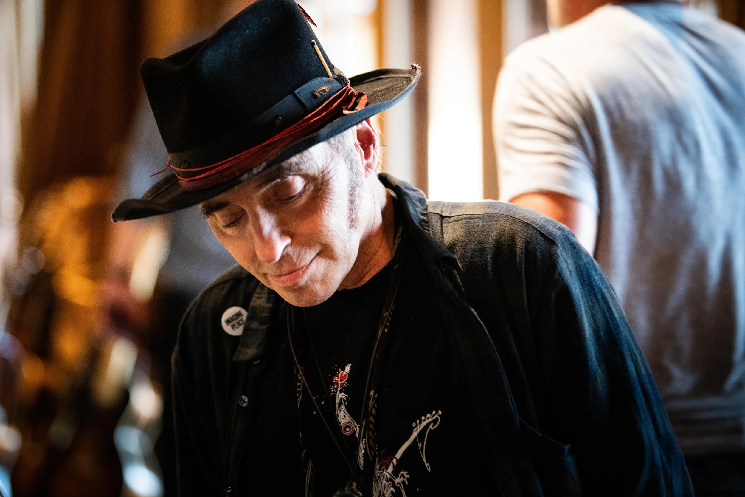 A man wearing a black hat with red accents, a button that says 'WHO THE,' a black shirt with a dragon design, and a black jacket, looking down indoors with a warm light background.