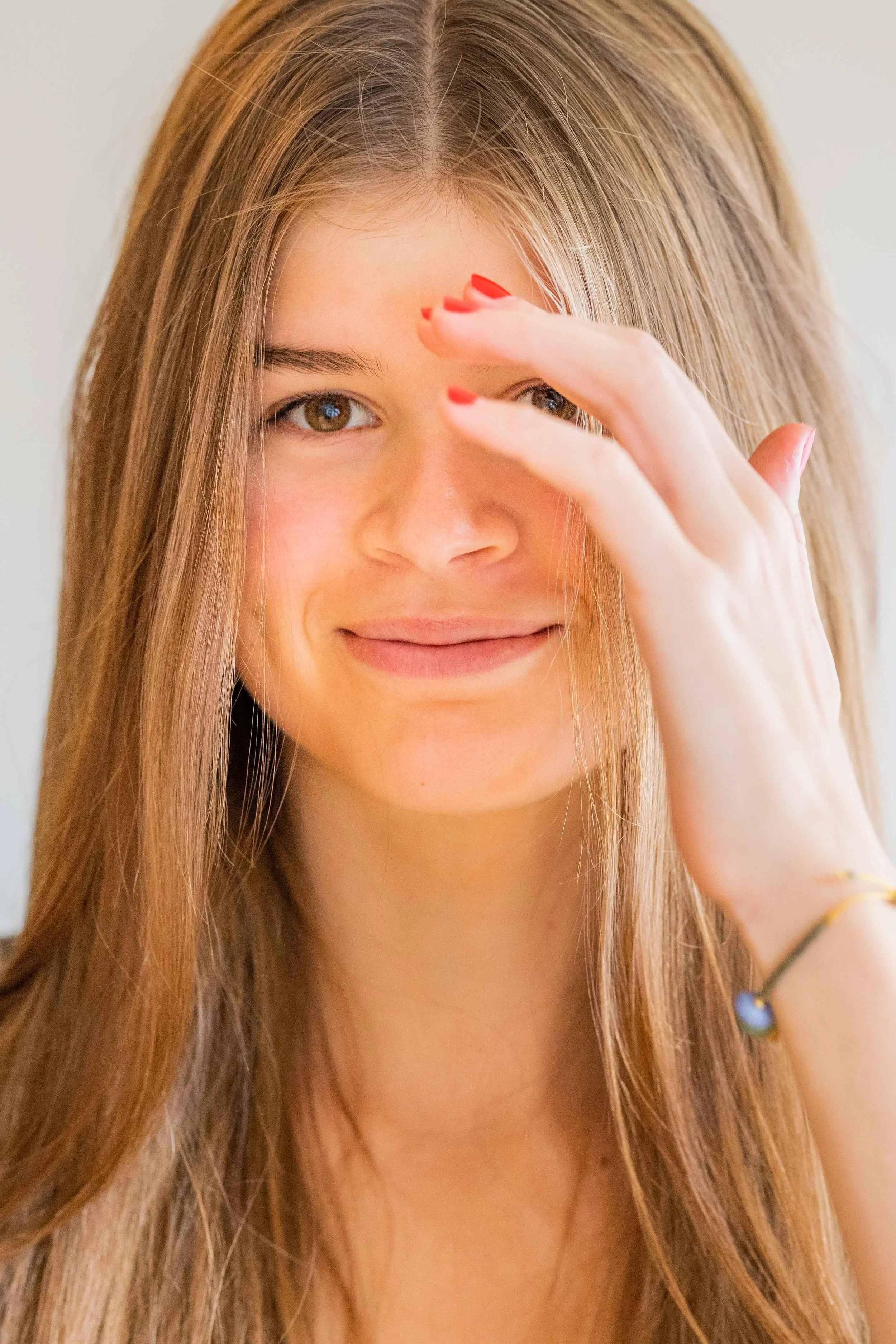 A young woman with long, light brown hair, smiling and winking, with one hand partially covering her face and painted red nails.