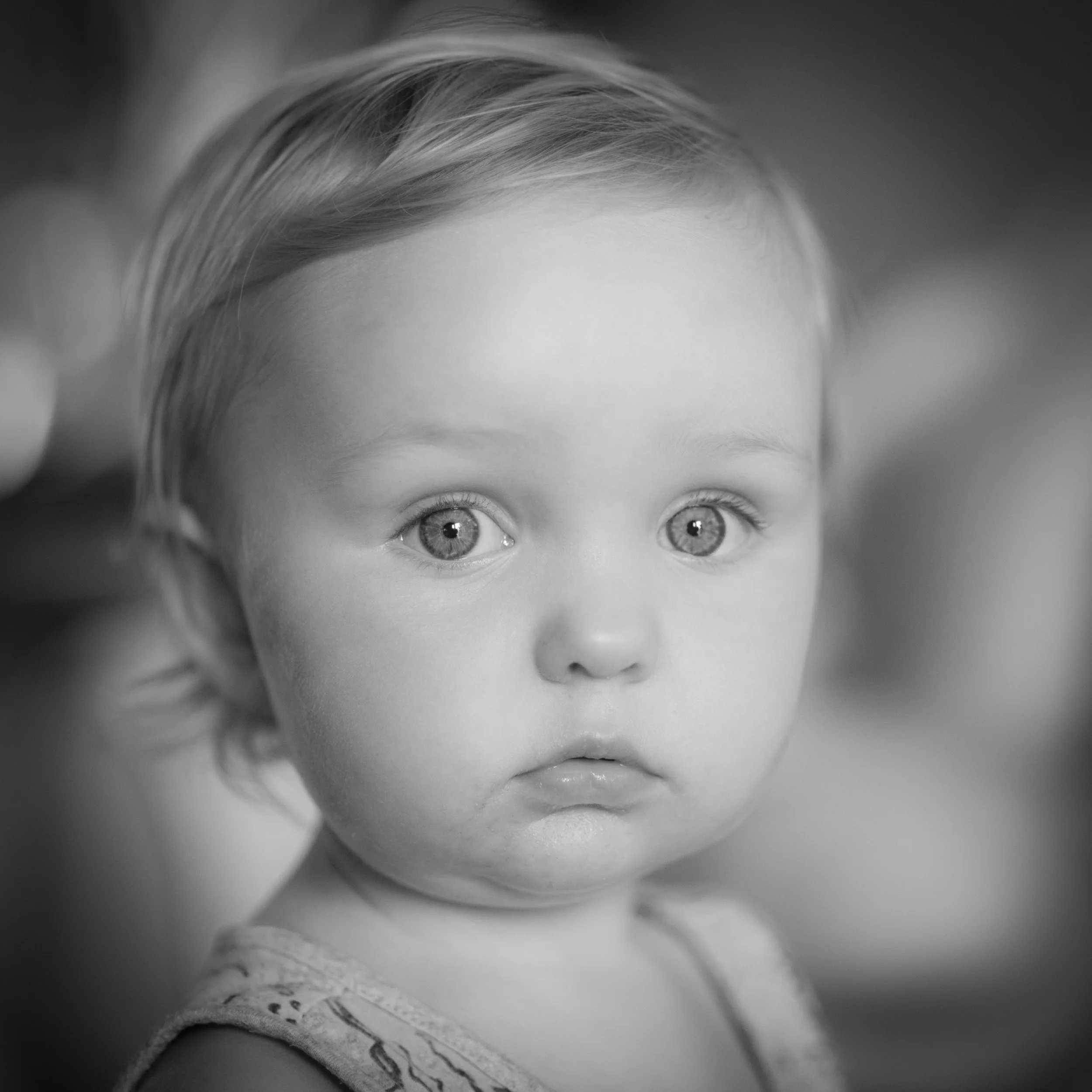 A black and white close-up photo of a young girl with big, light-colored eyes and light-colored hair, wearing a sleeveless top.