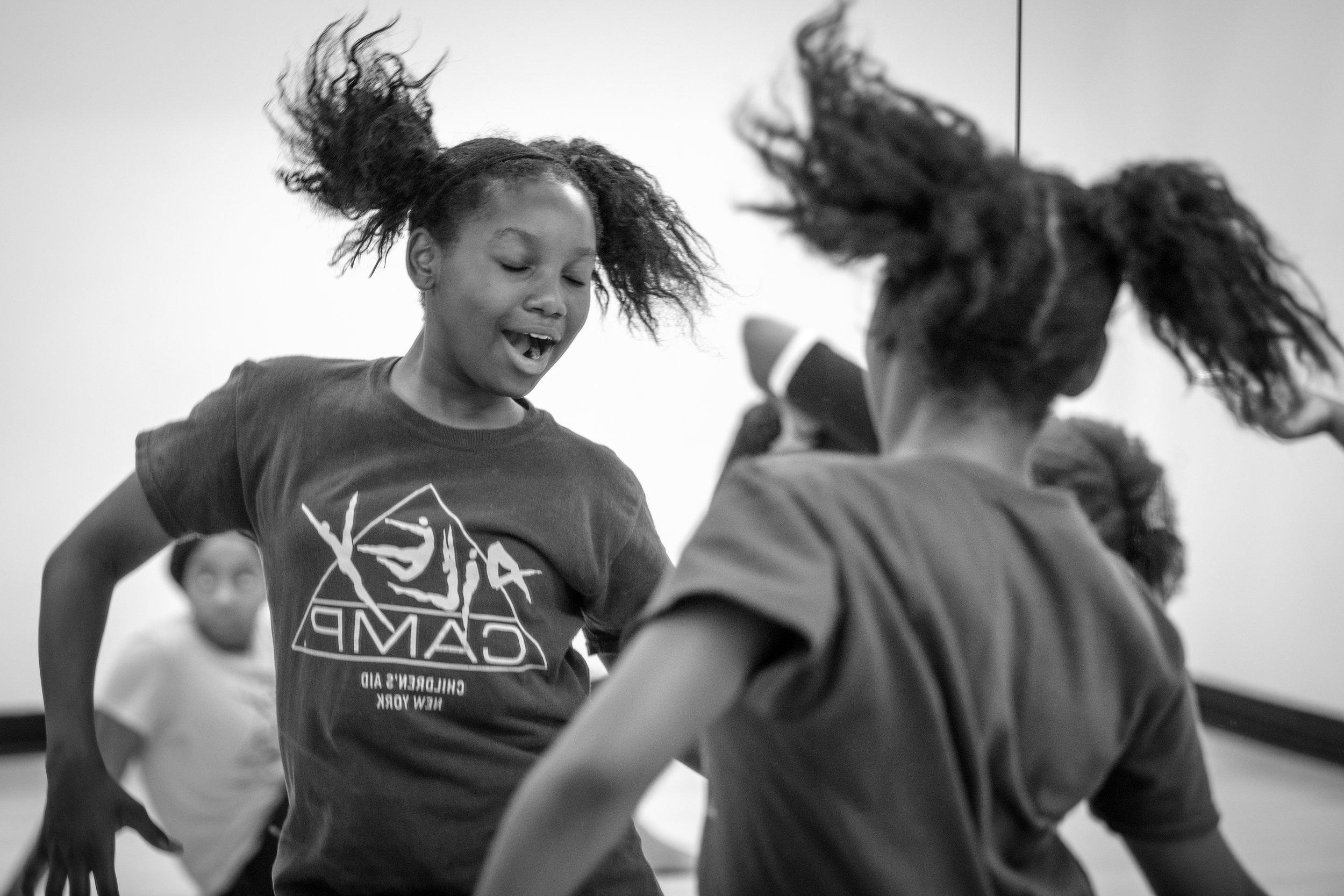 Two young girls with curly hair dancing and playing together in a room, with a third girl in the background.