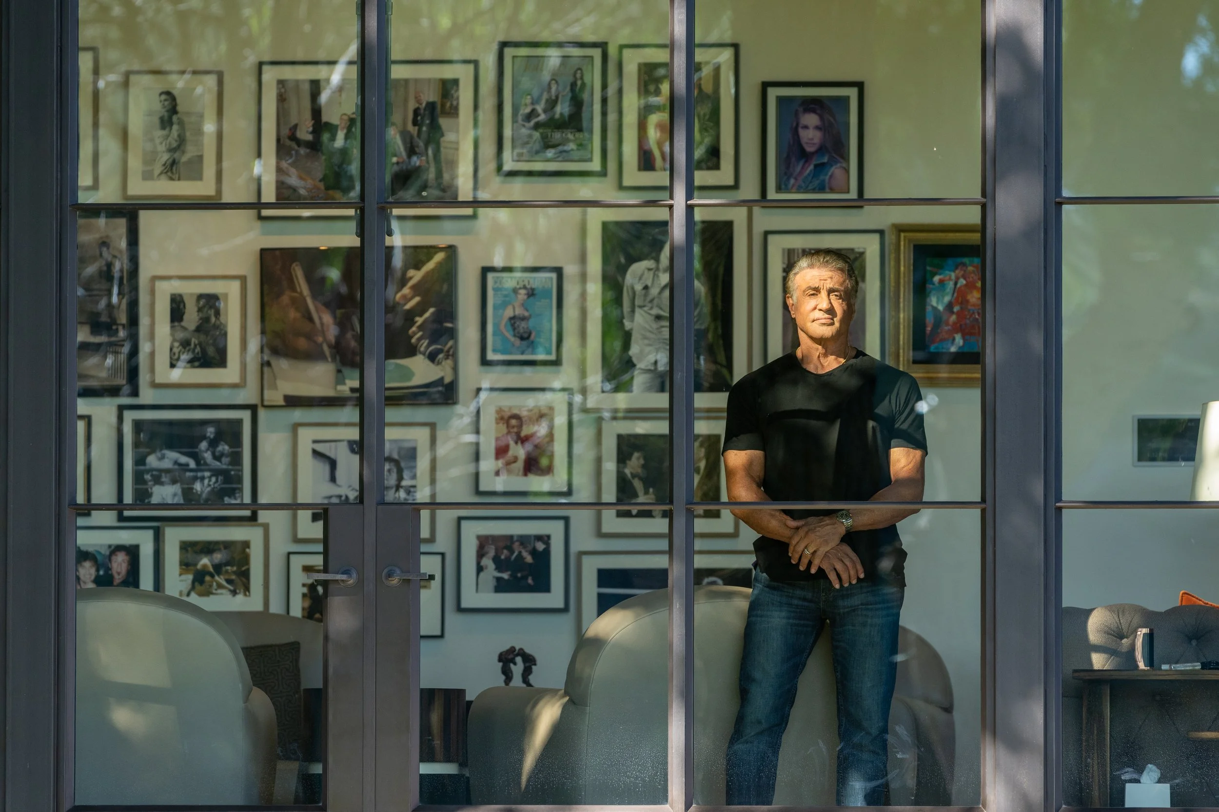 A man stands inside an art gallery, looking out through large glass windows. The gallery wall behind him features a variety of framed photographs and artwork.