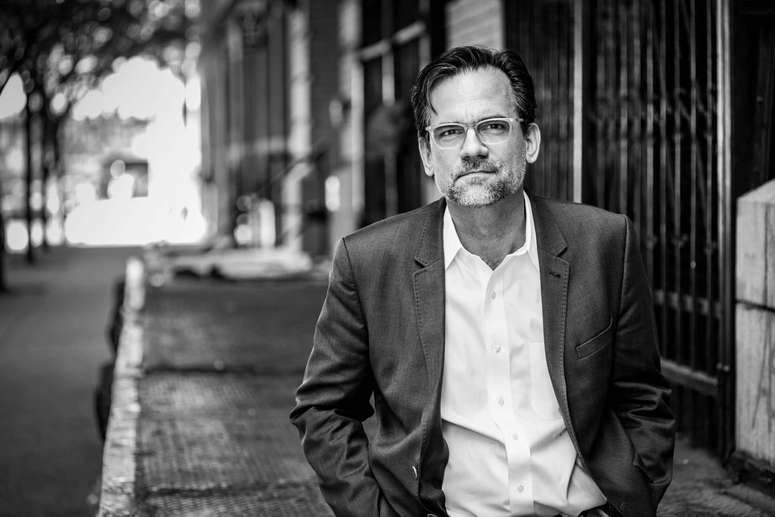 Black and white photo of a middle-aged man with glasses, wearing a suit and white shirt, standing on a city sidewalk with cars and trees in the background.