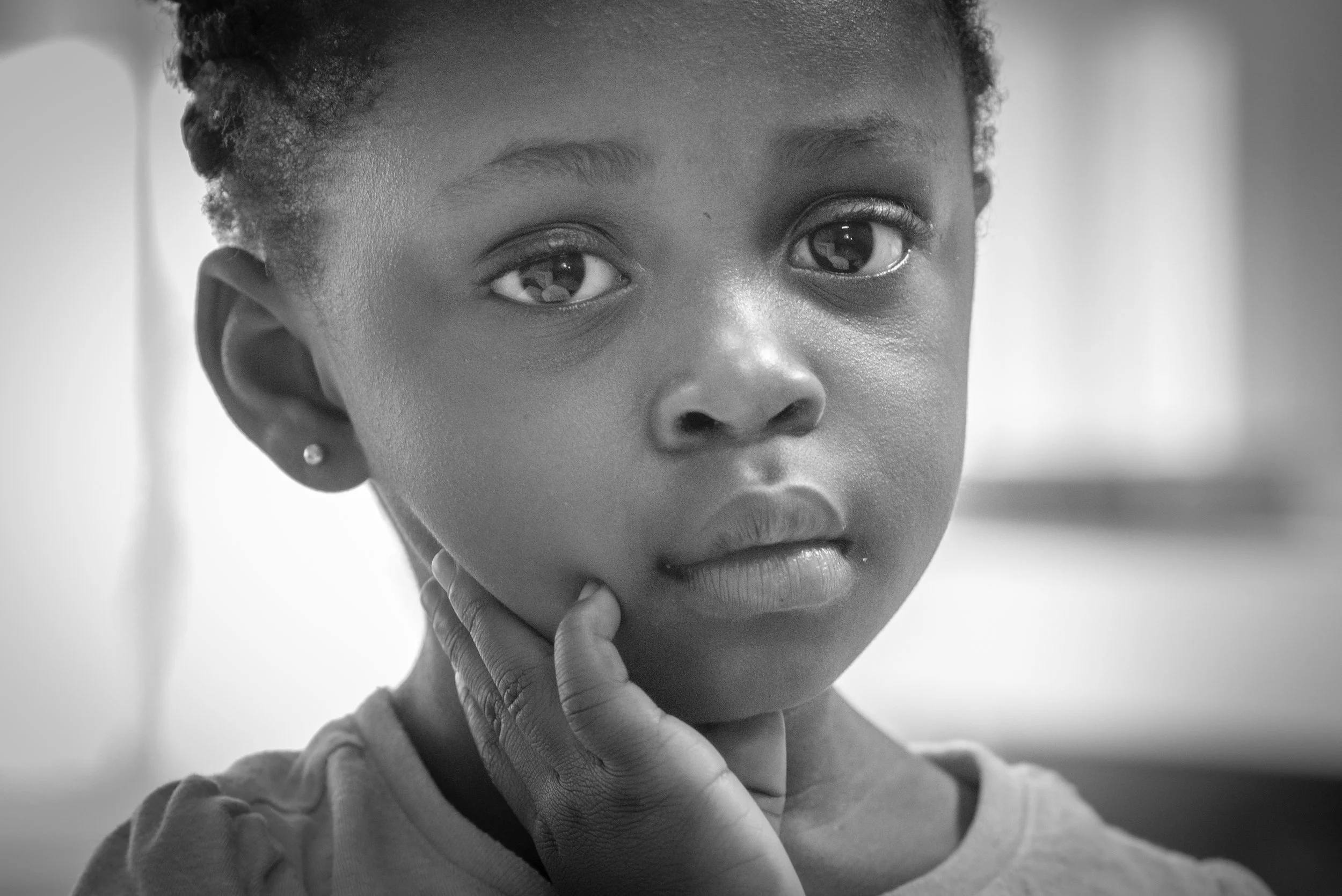 A young girl with short curly hair and earrings, looking directly at the camera with her hand touching her face, in black and white.