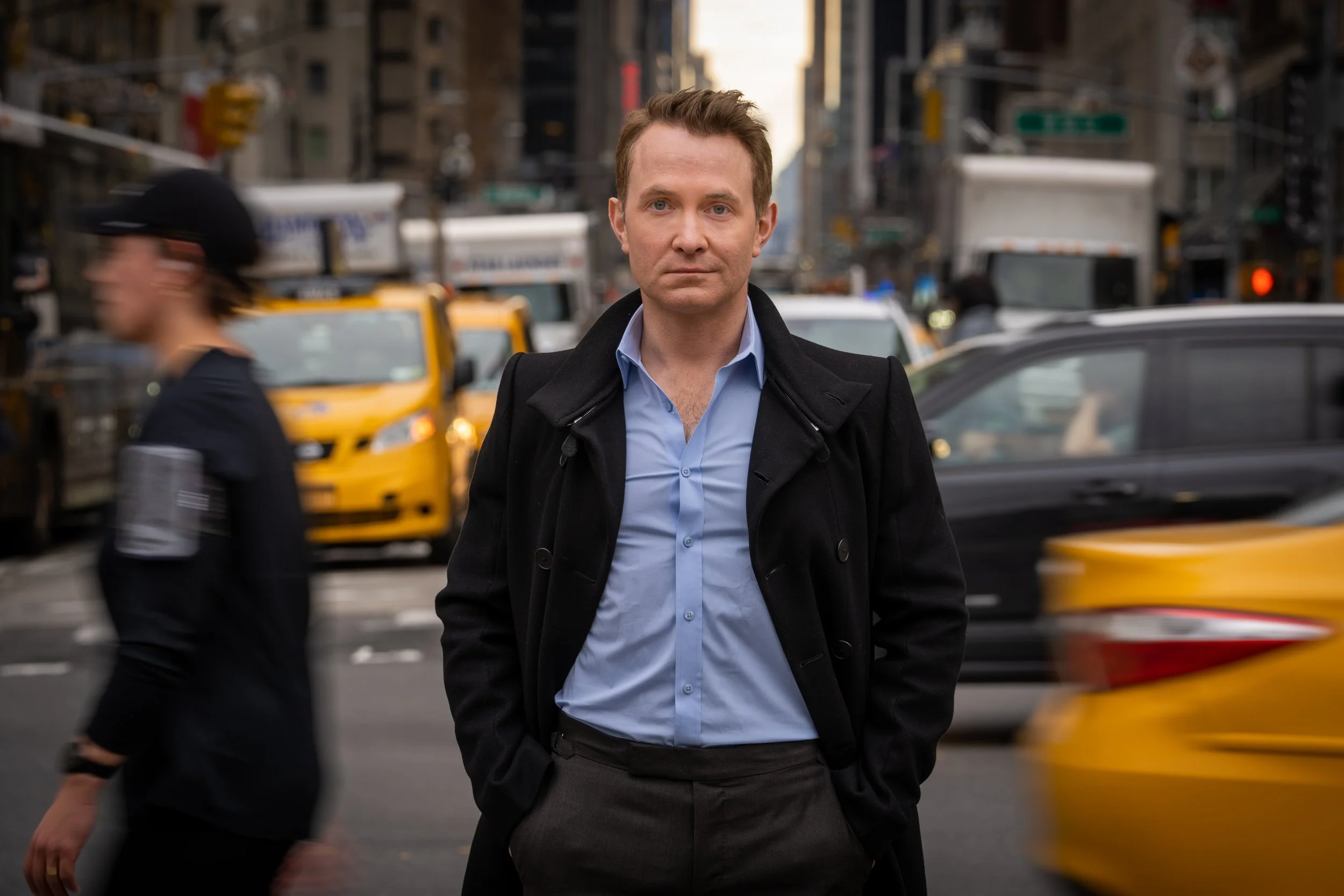 A man in a black coat and blue shirt standing in the street with yellow taxis and blurred city traffic in the background.