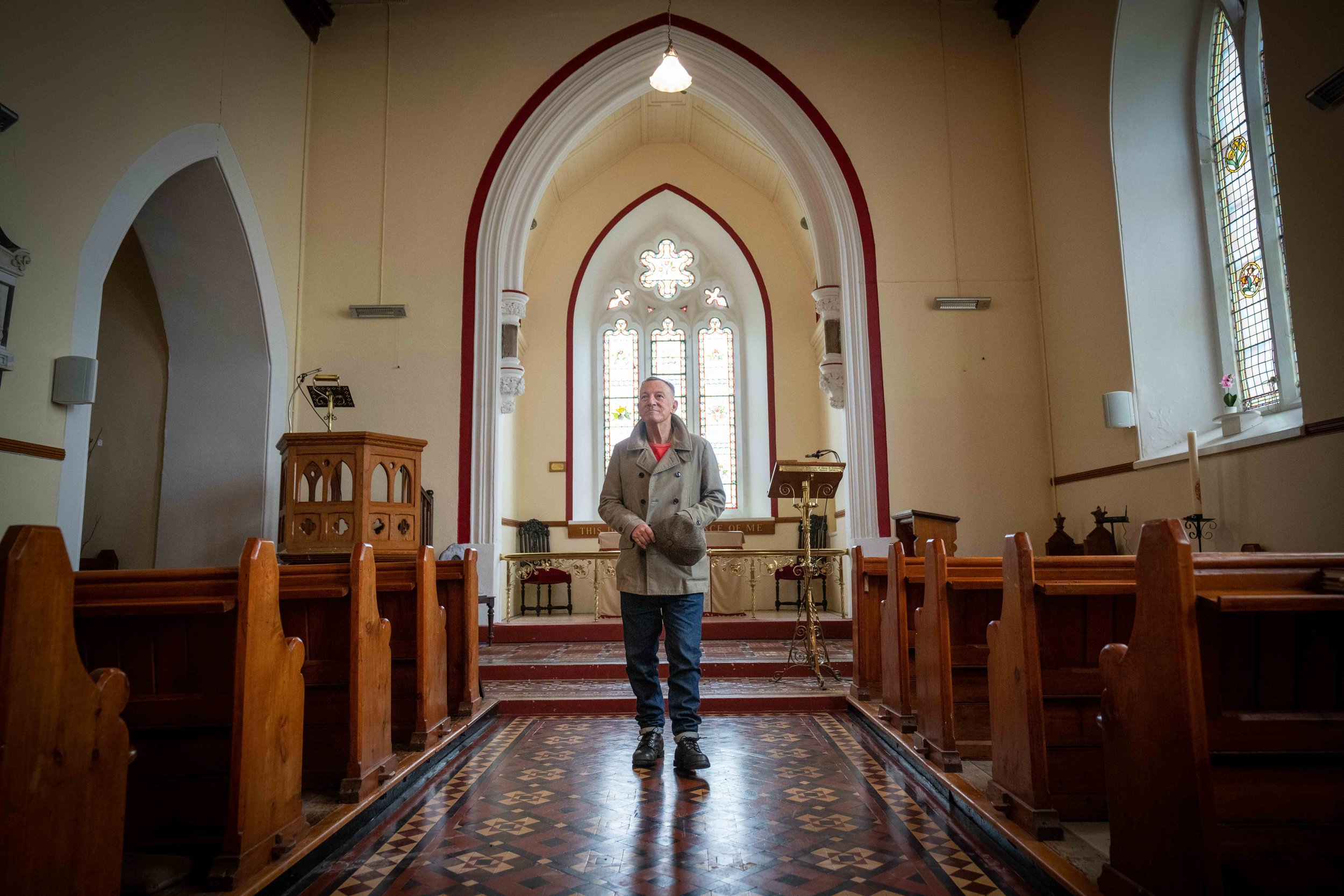 A man standing inside a church near the altar, looking up. The church has wooden pews, stained glass windows, and decorative architectural features.