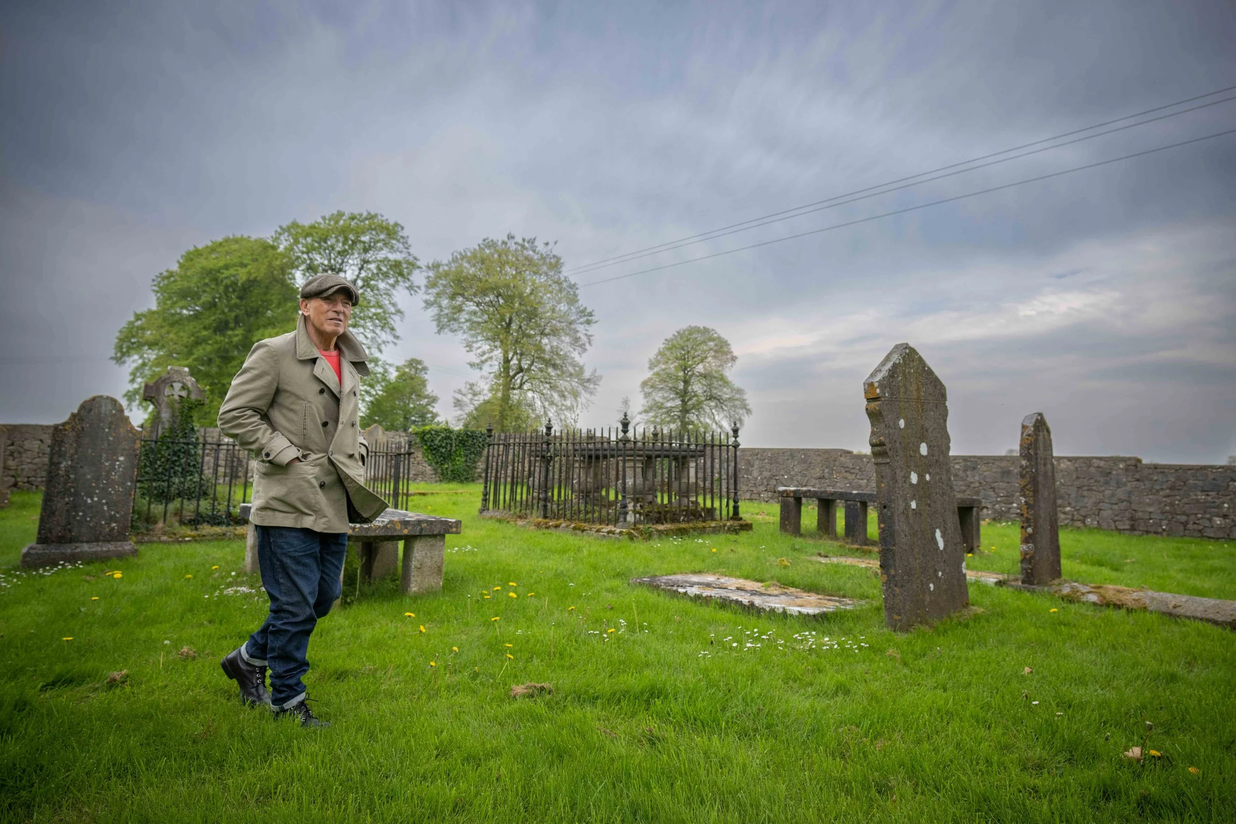 A man wearing a tan trench coat and a gray cap standing in a grassy graveyard with old tombstones and a stone wall in the background, under a cloudy sky.