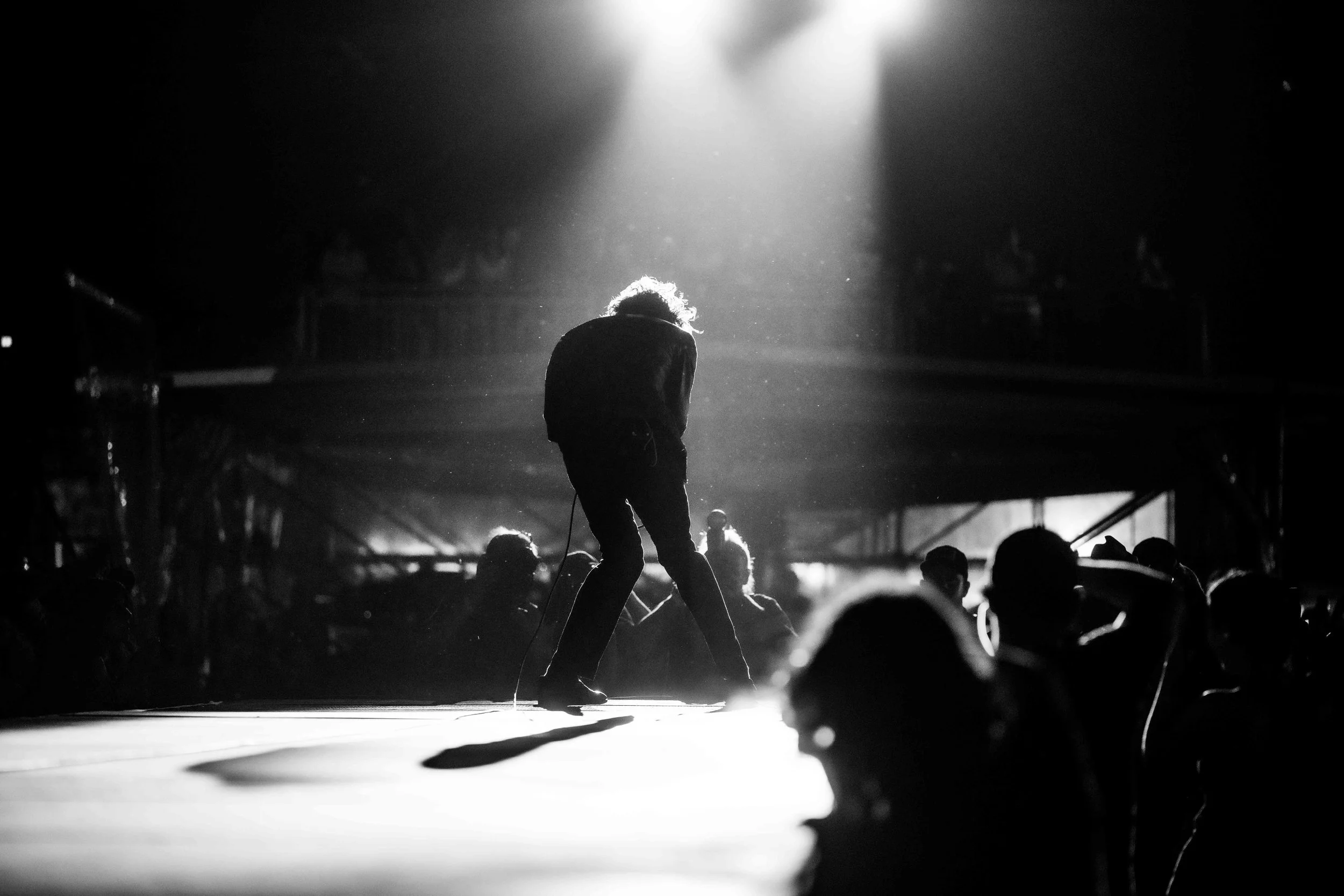 Black and white image of a person crouched on stage with a microphone cable, backlit by a bright spotlight, with an audience in the background.