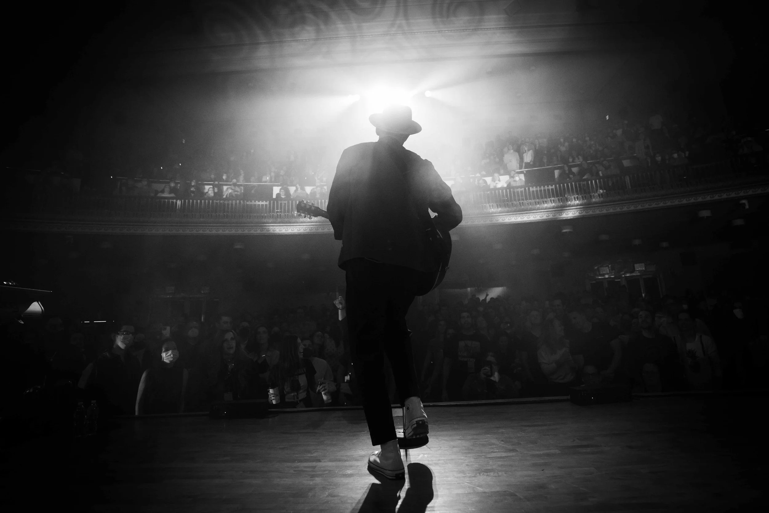 A musician in a hat playing guitar on stage in front of an audience, with bright backlighting creating a silhouette effect.