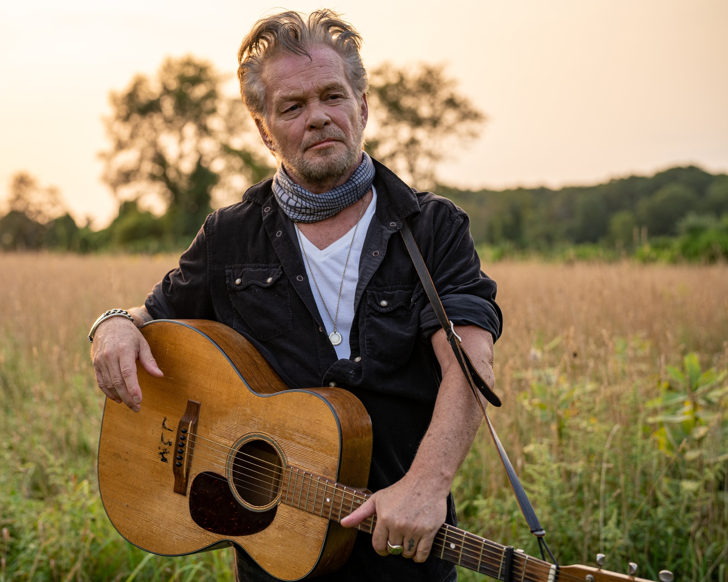 A man with gray hair and a beard playing an acoustic guitar outdoors during sunset, standing in a grassy field with trees in the background.