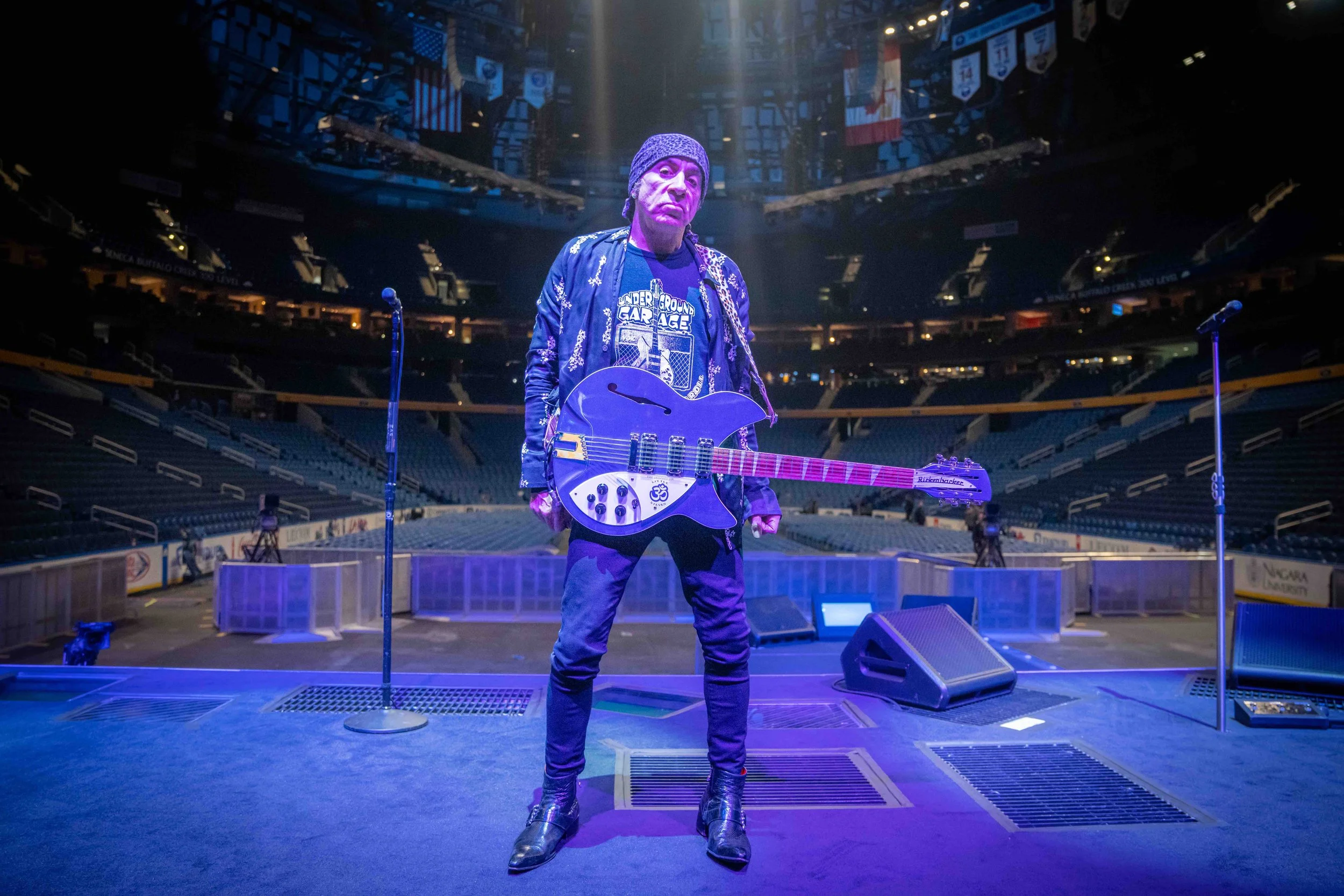 A man standing on a music stage holding a guitar, with microphone stands on either side of him, in an empty arena with colored stage lighting.