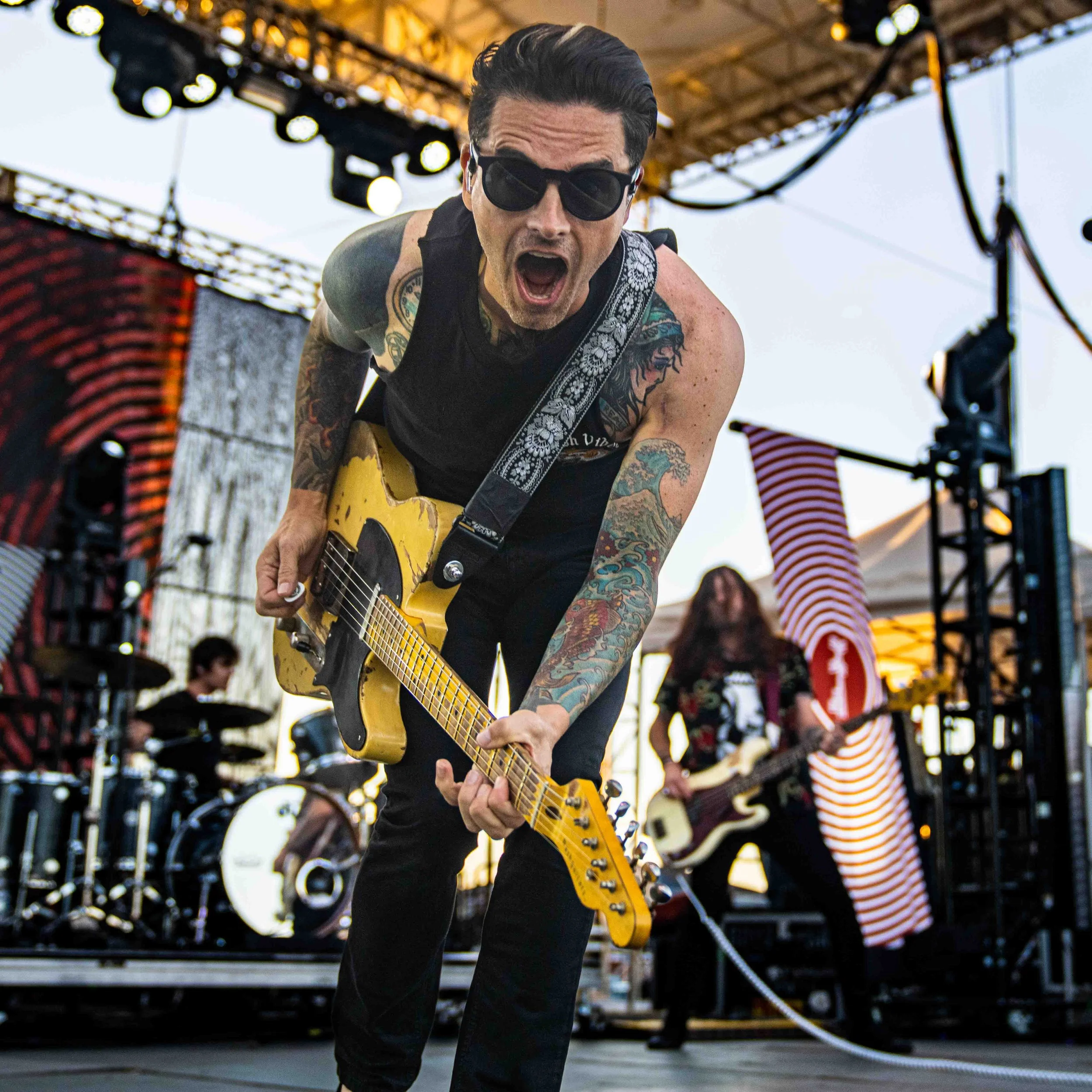 A male guitarist with sunglasses and tattoos, passionately playing a yellow electric guitar on stage during a live outdoor concert, with band members and stage equipment in the background.