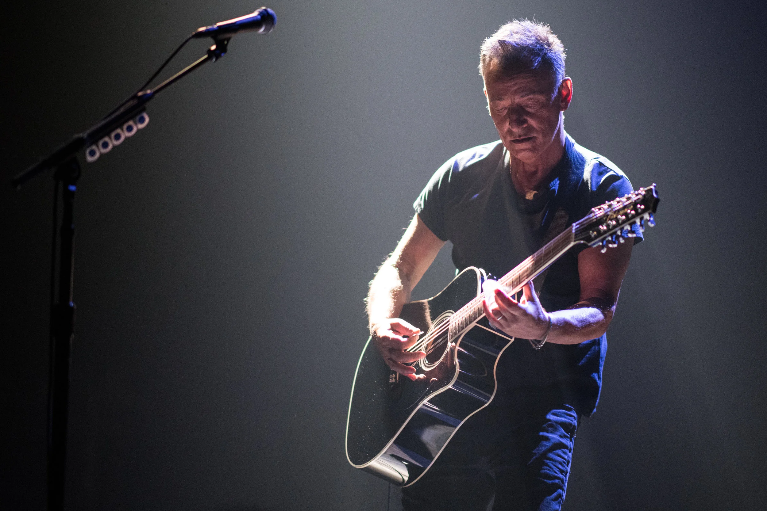 A man playing an acoustic guitar on stage under a spotlight with a microphone in front.
