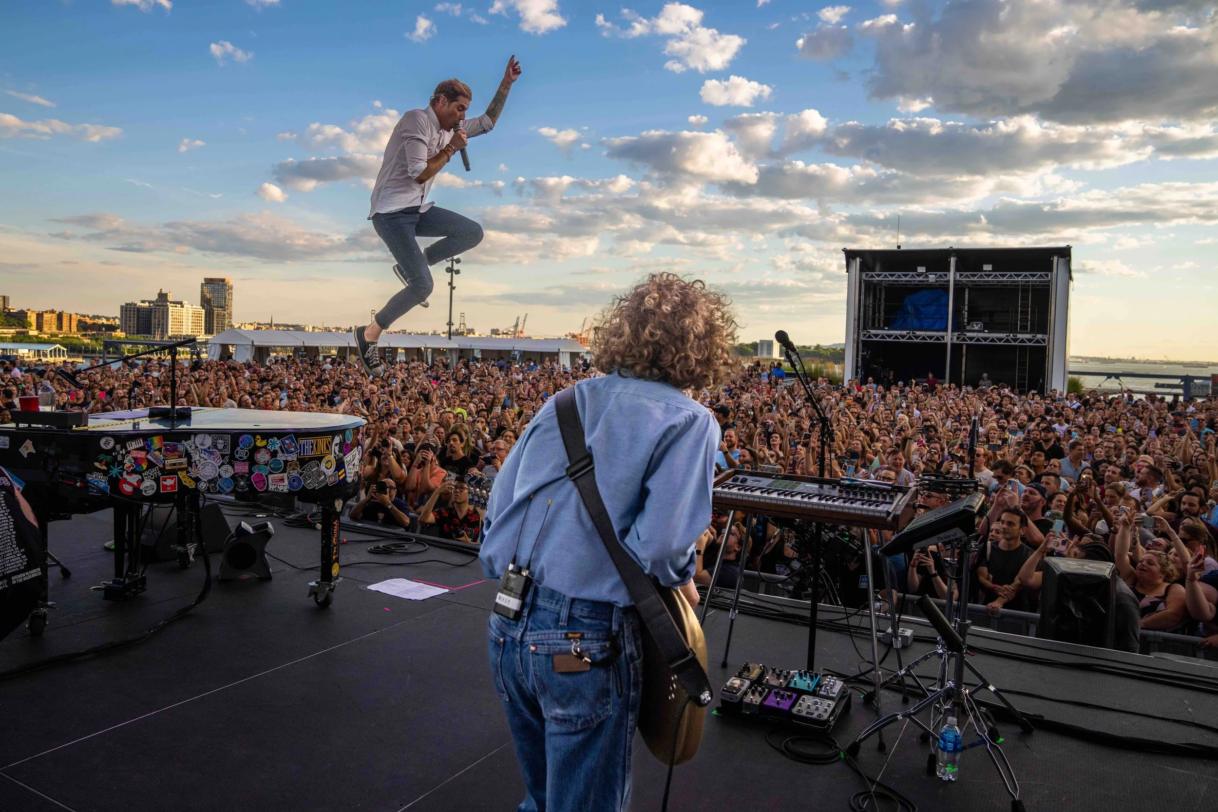 A live outdoor concert showing a lead singer jumping with a microphone, and a guitarist facing the stage, with a large crowd of concertgoers, city skyline, and cloudy sky in the background.