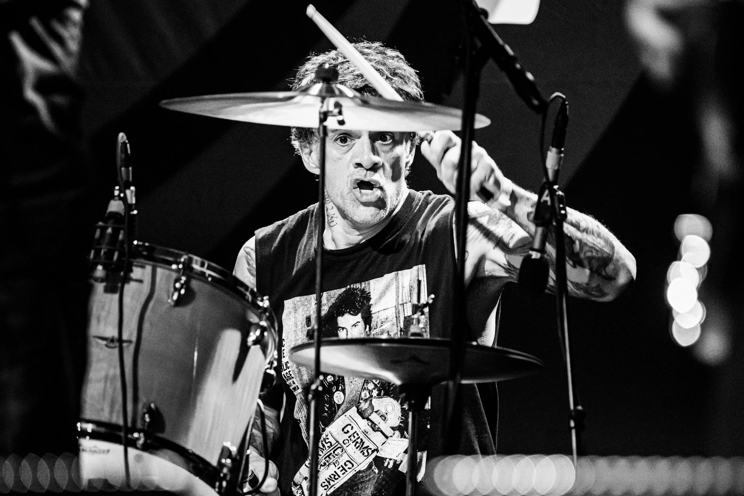 Black and white photo of a drummer with curly hair, tattoos, and a sleeveless shirt playing drums on stage, with an intense expression.