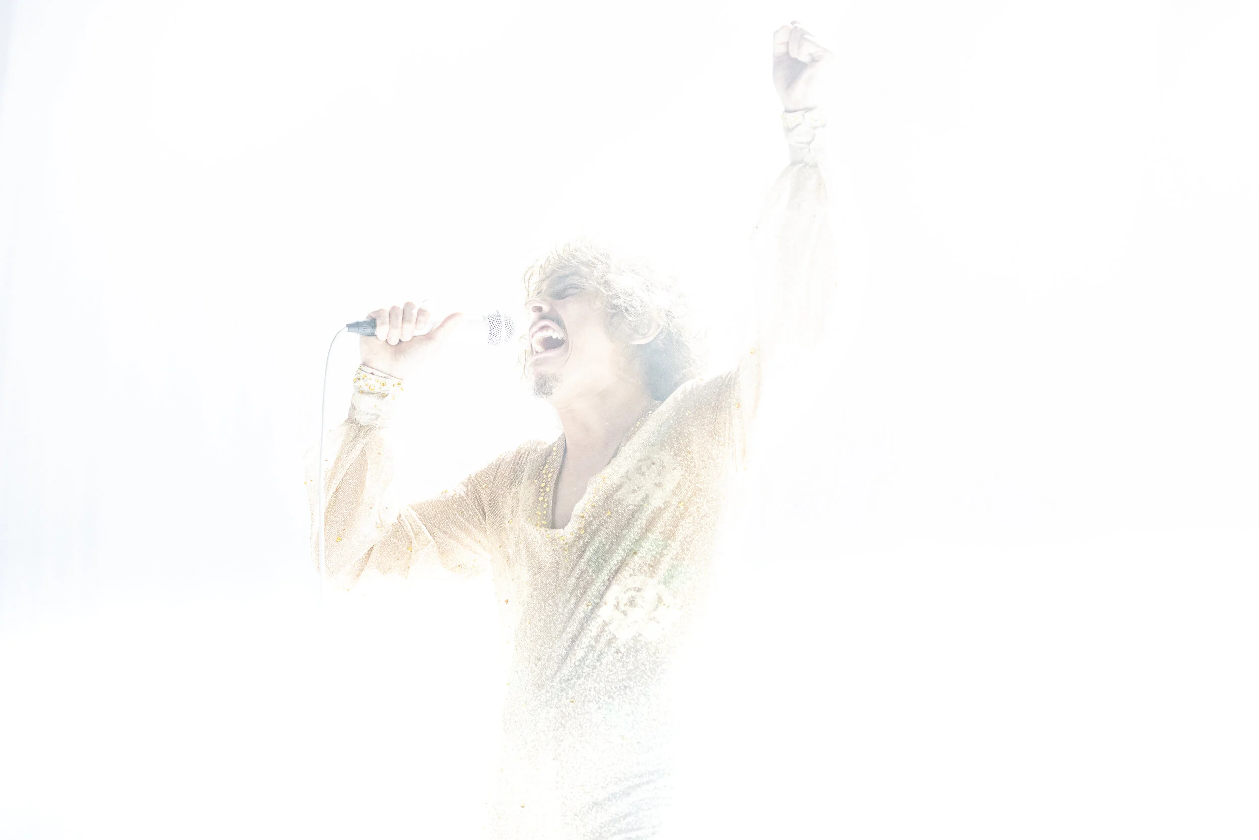 A person singing into a microphone with a passionate expression, raised fist, and wearing a glittery gold outfit, surrounded by bright, white light.