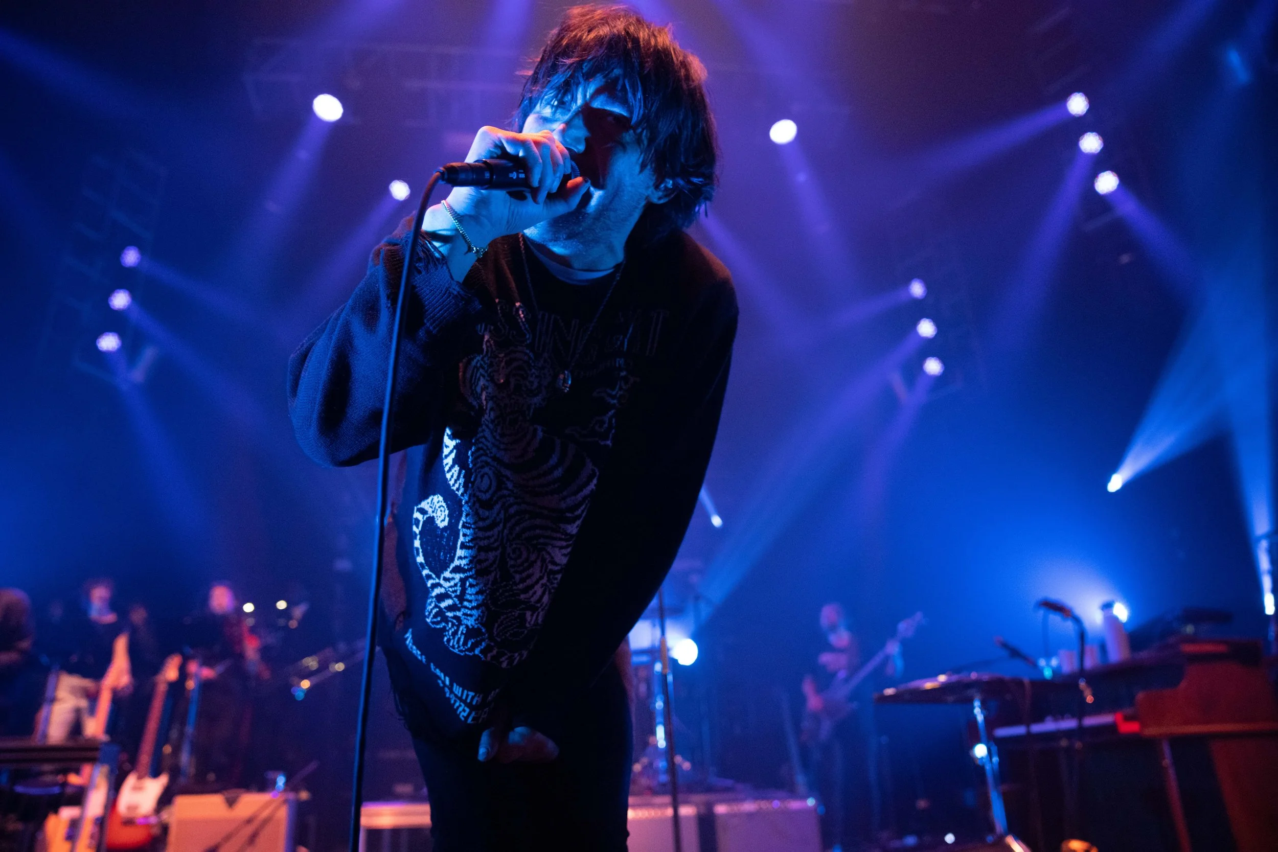 A male singer with shaggy hair and sunglasses performs with a microphone on stage, surrounded by blue and purple stage lighting, with band members and musical instruments in the background.