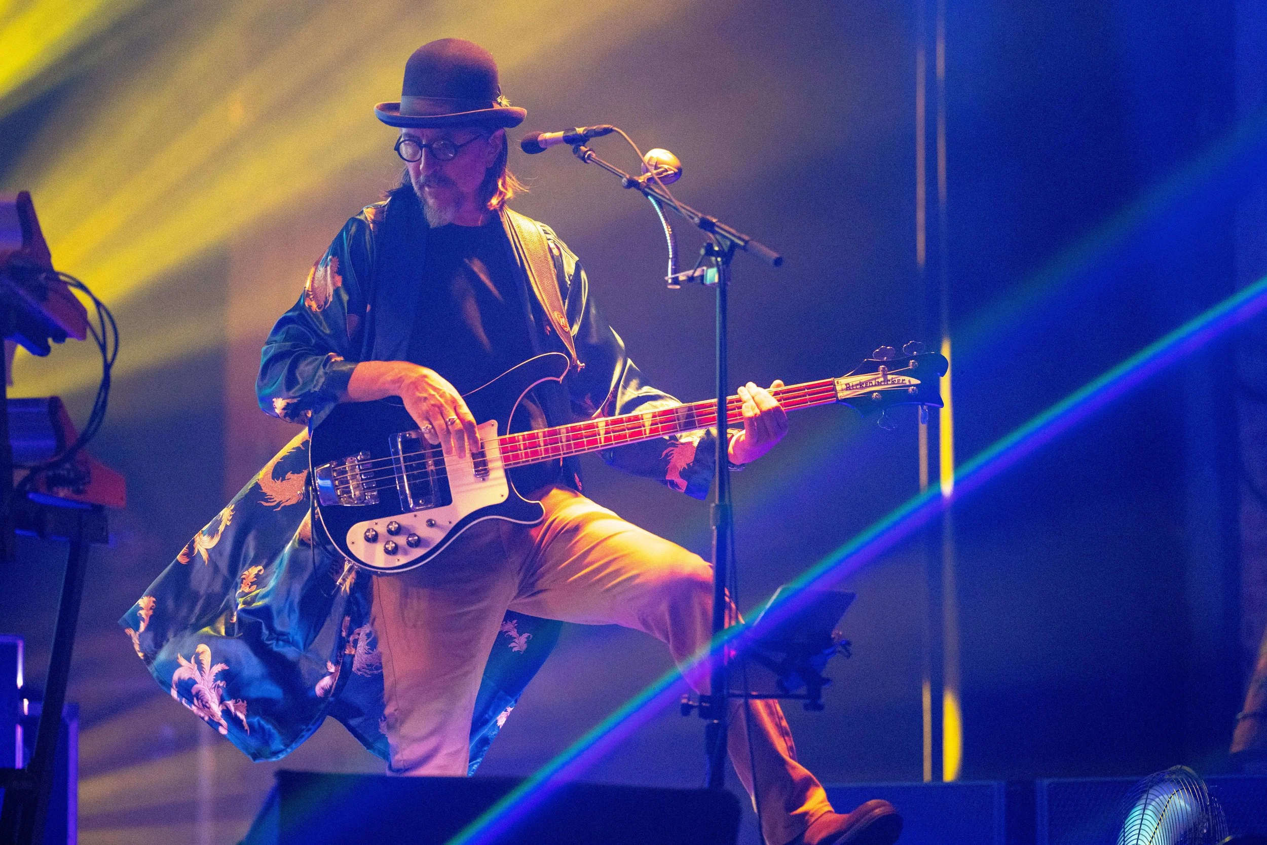 A musician wearing a black hat, glasses, and a floral kimono-style jacket plays a bass guitar on stage with colorful lighting and fog effects.