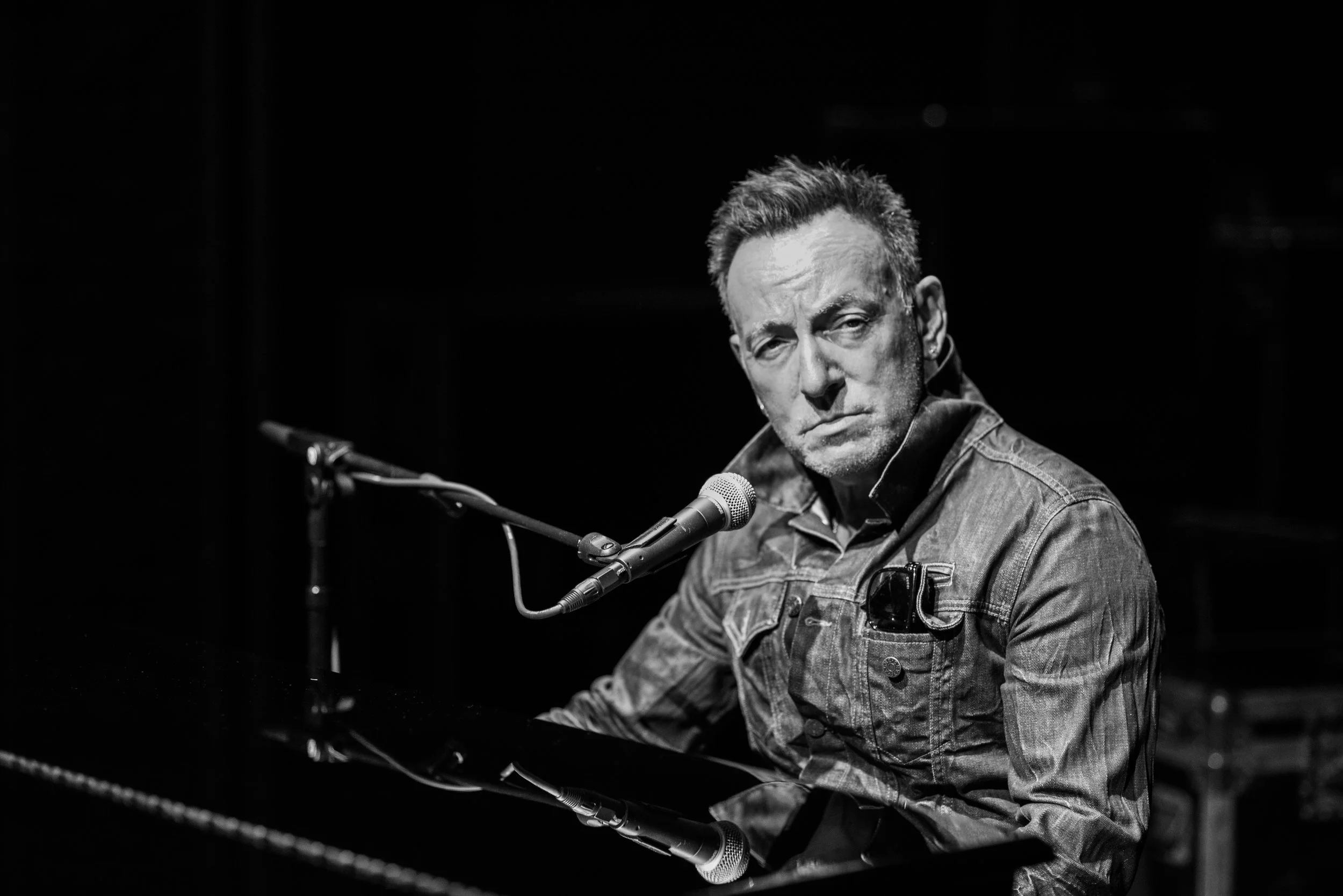 Black and white photo of a man with short hair and a serious expression, sitting at a piano with a microphone, wearing a denim jacket.