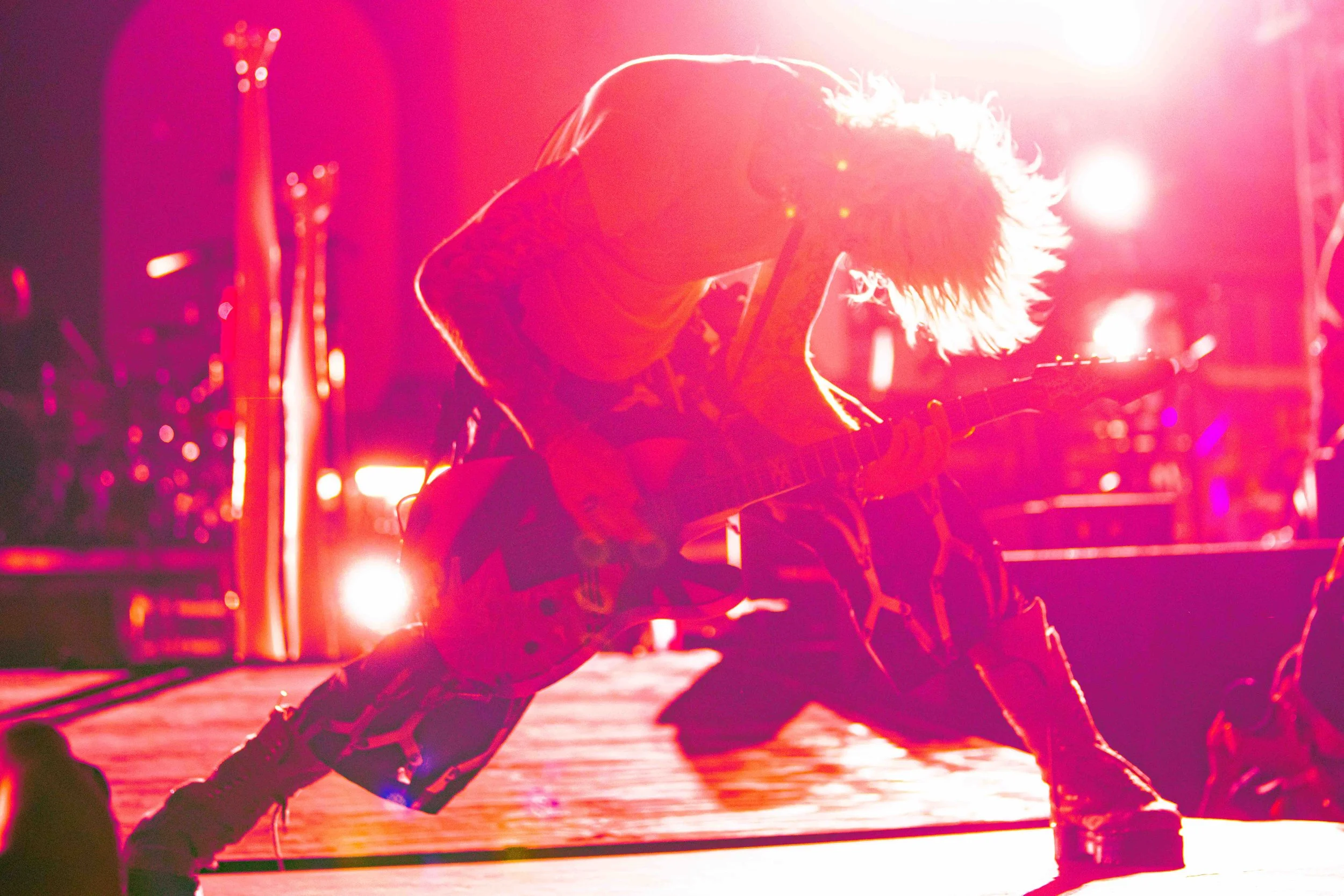 A musician playing an electric guitar on stage in front of bright pink and purple lighting, with hair flying as they perform energetically.