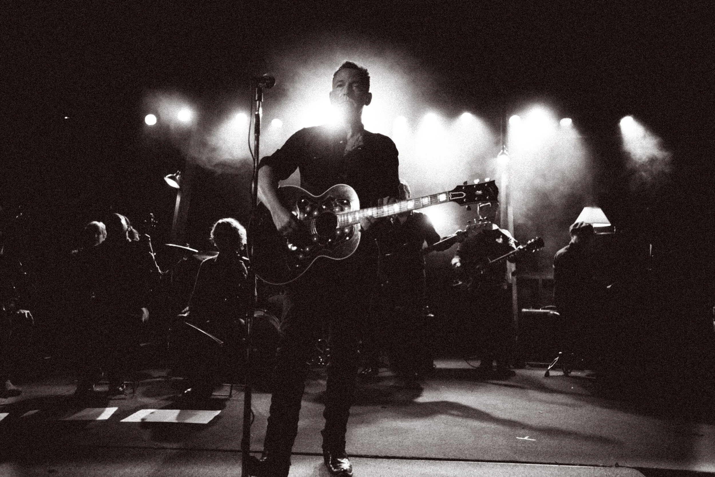 A musician playing an electric guitar on stage, backlit with bright lights, surrounded by a group of seated musicians in a dark venue.