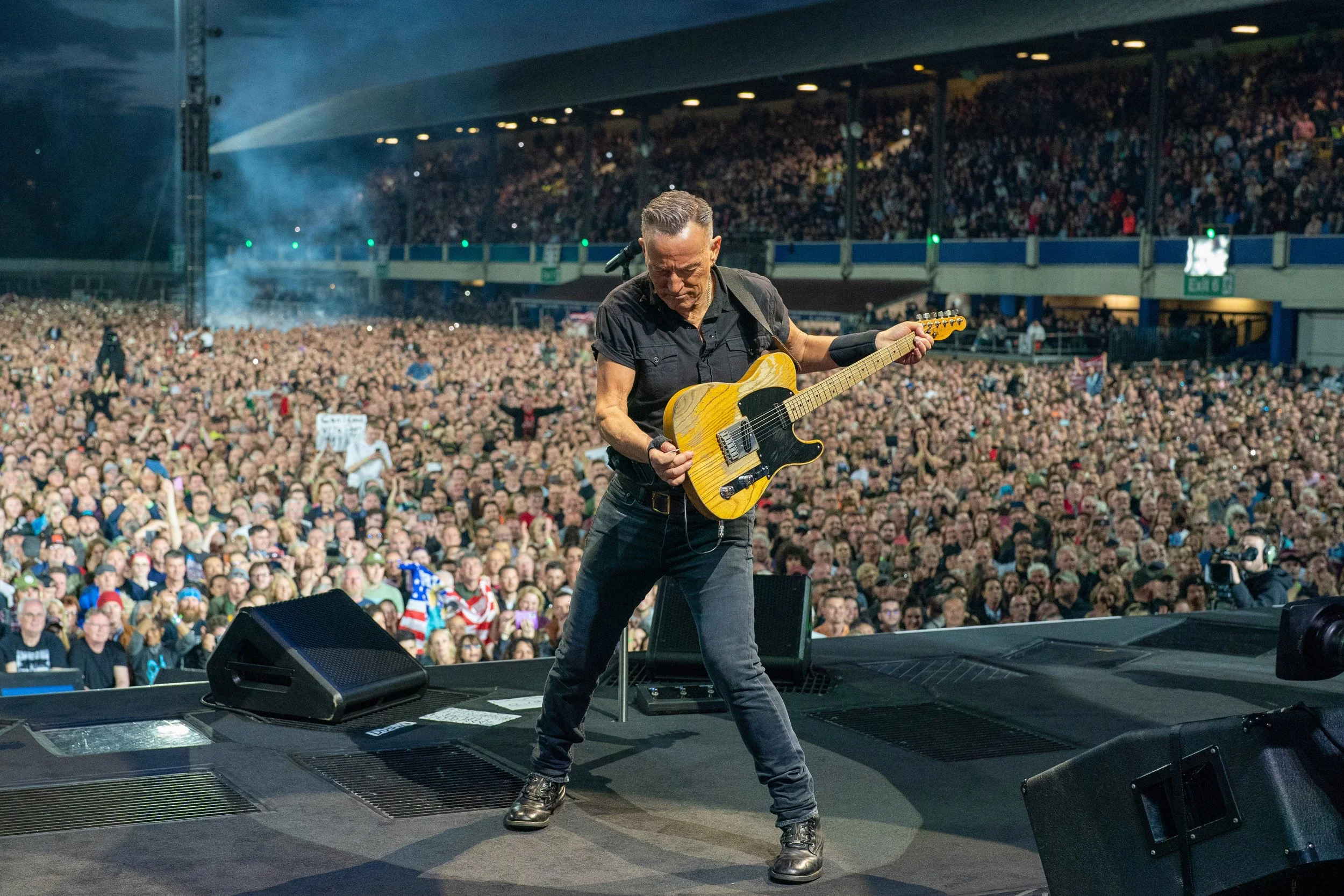 Male guitarist performing on stage with a large crowd in the background at a concert.