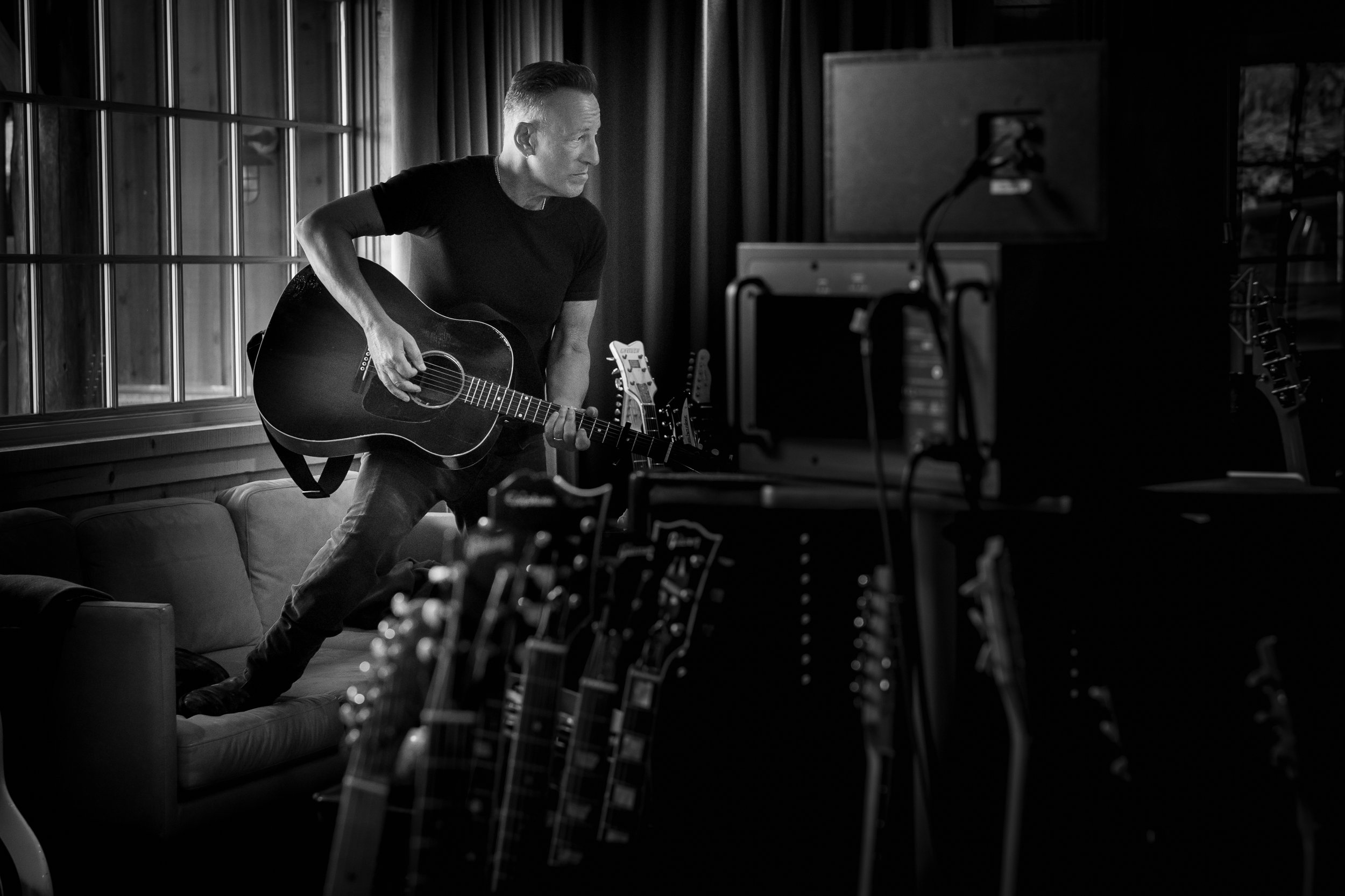 A man in a black t-shirt and jeans playing an acoustic guitar in a room with large windows and multiple guitars in the foreground, in black and white.