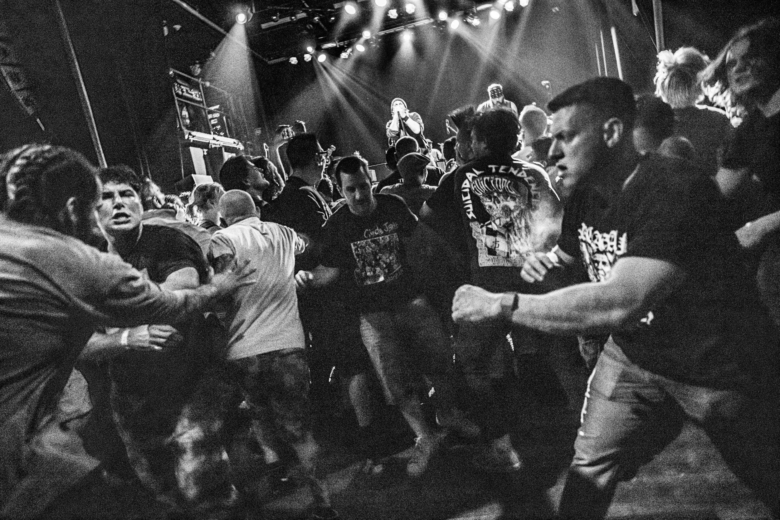 Black and white photo of people crowding and fighting in front of a stage at a concert, with a performer singing into a microphone under bright stage lights.
