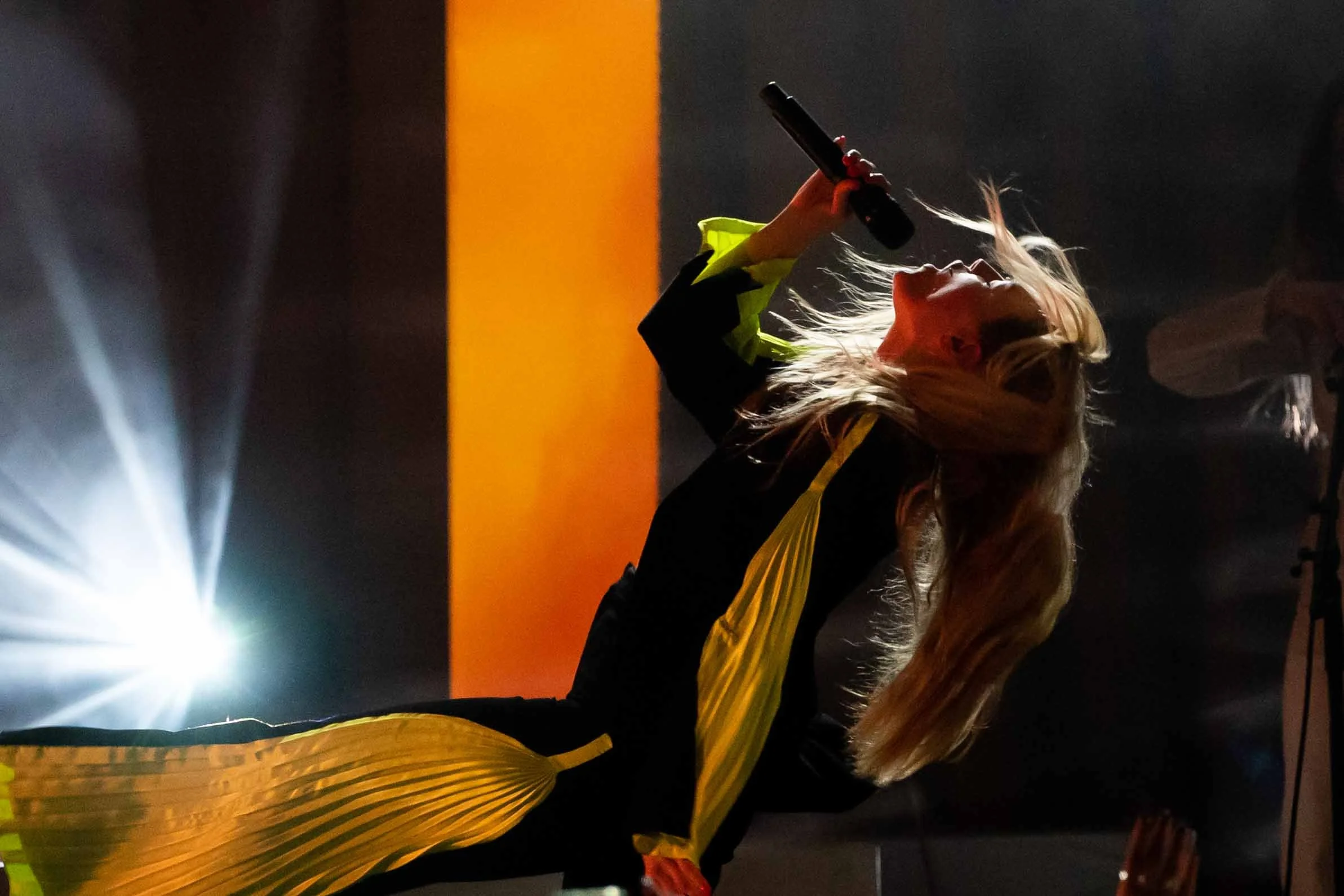 Female singer performing on stage, holding a microphone, with colorful stage lighting and a dark background.