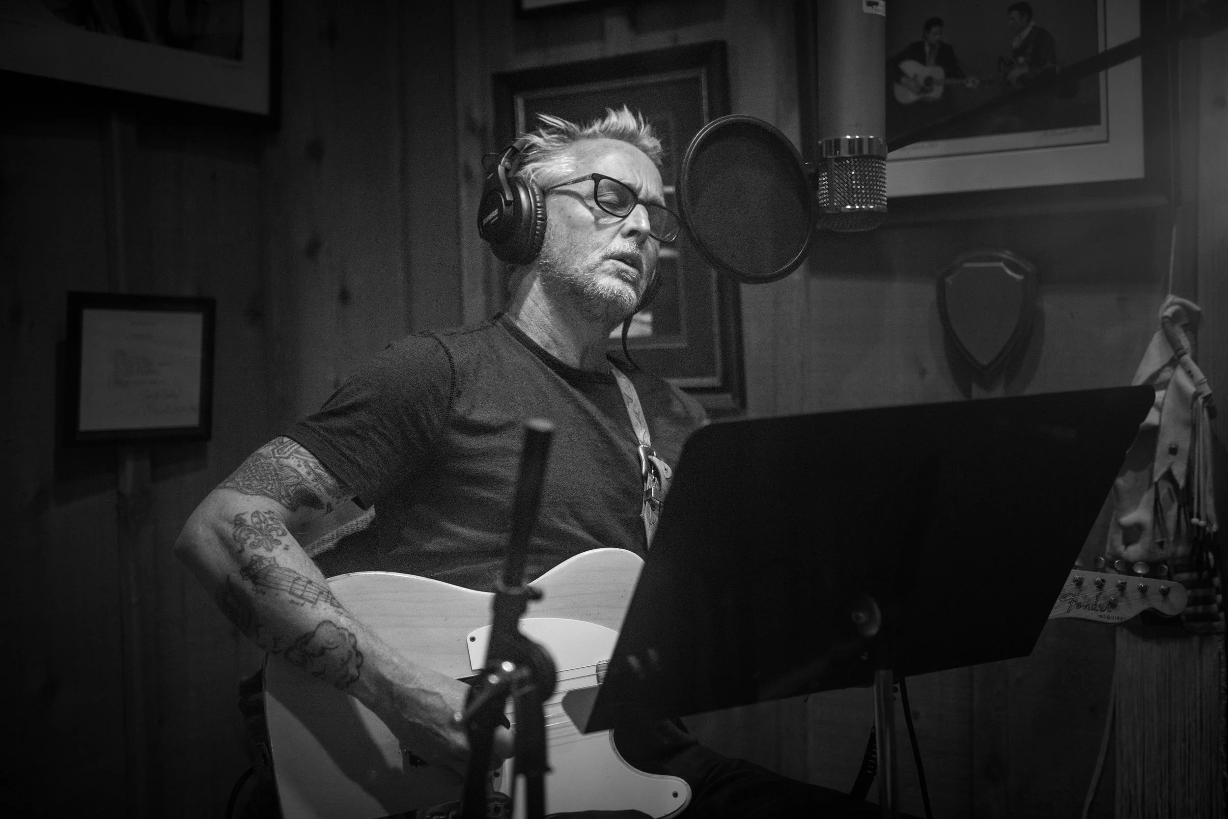 A man with glasses, tattoos, and gray hair wearing headphones, singing in a recording studio while playing an acoustic guitar.