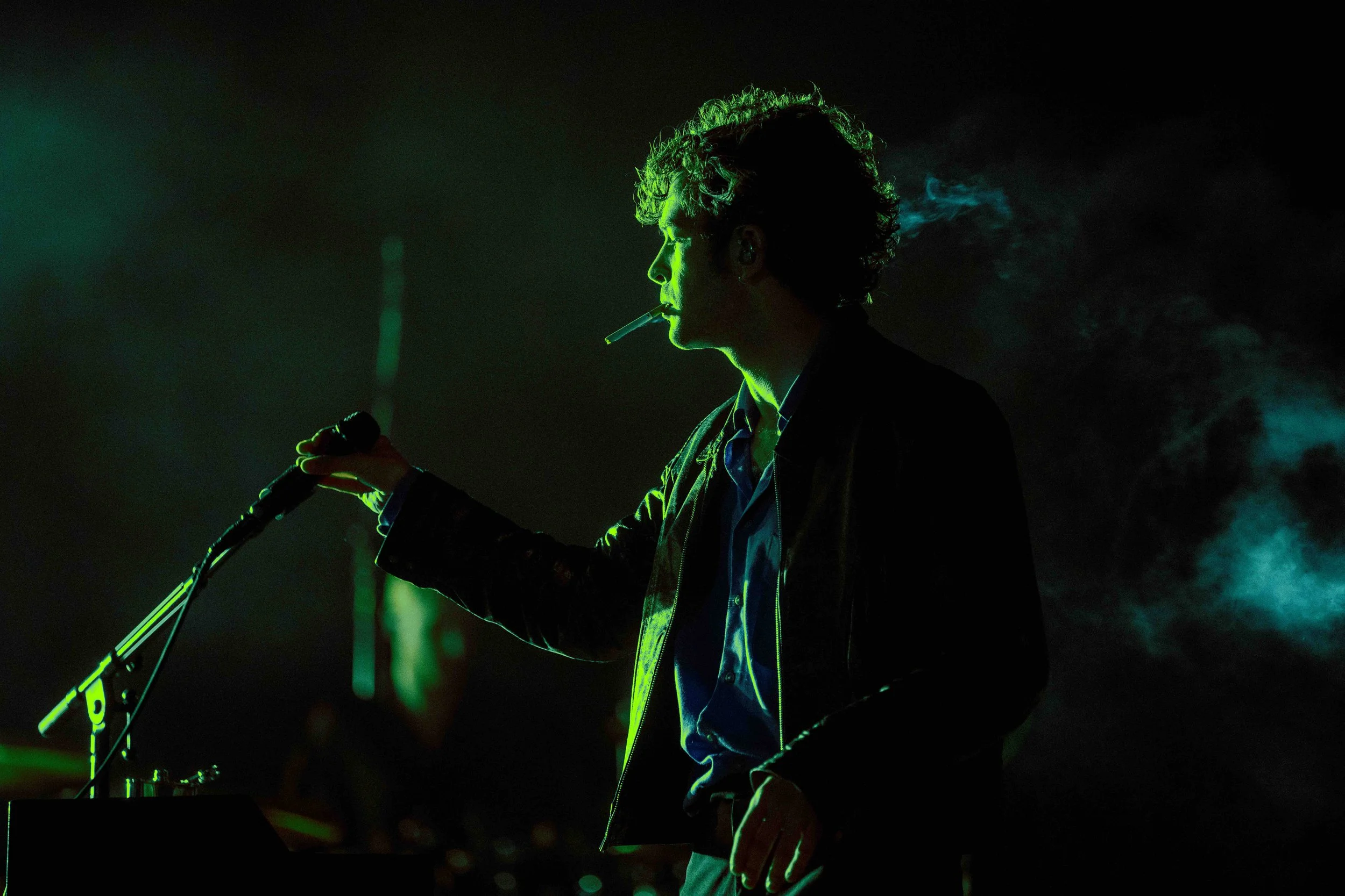 Silhouette of a person standing at a microphone with a cigarette in their mouth, illuminated by green light and surrounded by smoke.