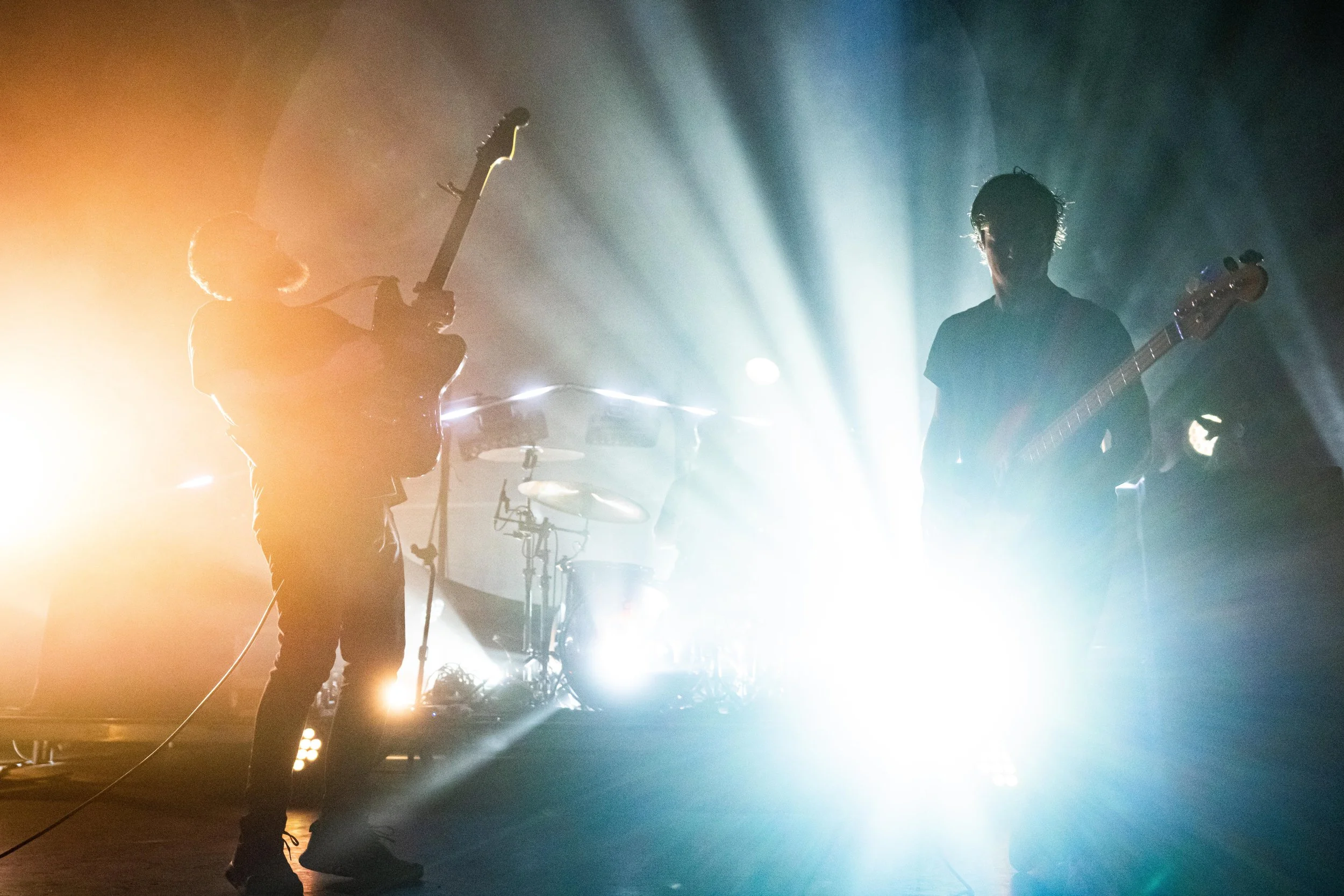 Silhouettes of two musicians playing guitars on stage with bright stage lights and fog effects.