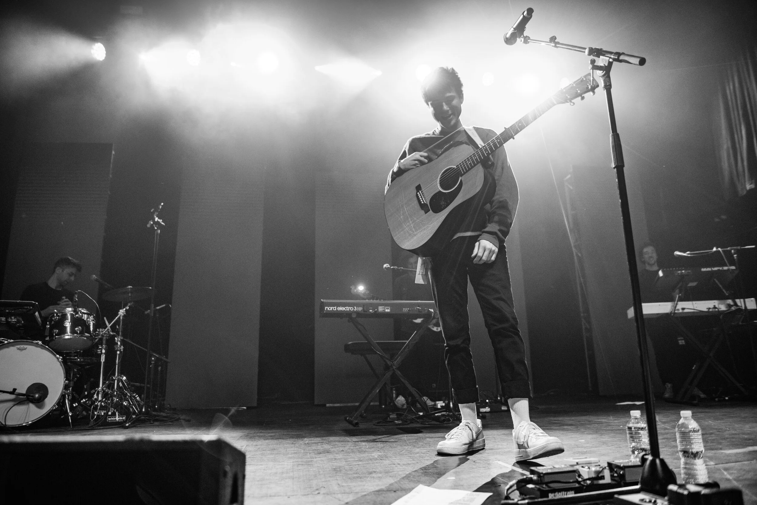 A musician on stage holding an acoustic guitar, smiling, with a keyboard and drums in the background and stage lights overhead.