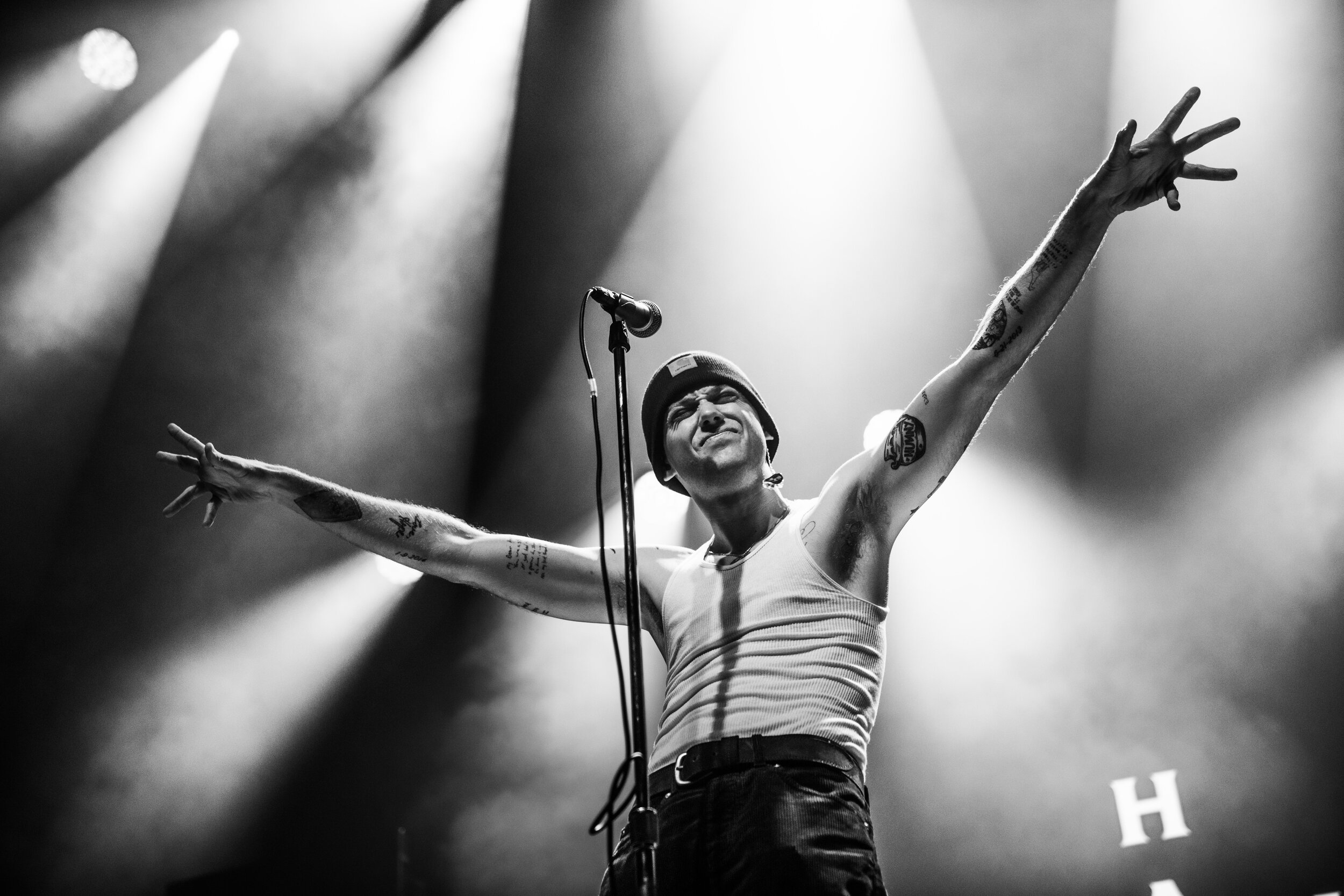 A performer on stage with arms outstretched, wearing a beanie, tank top, and displaying tattoos, under stage lighting.