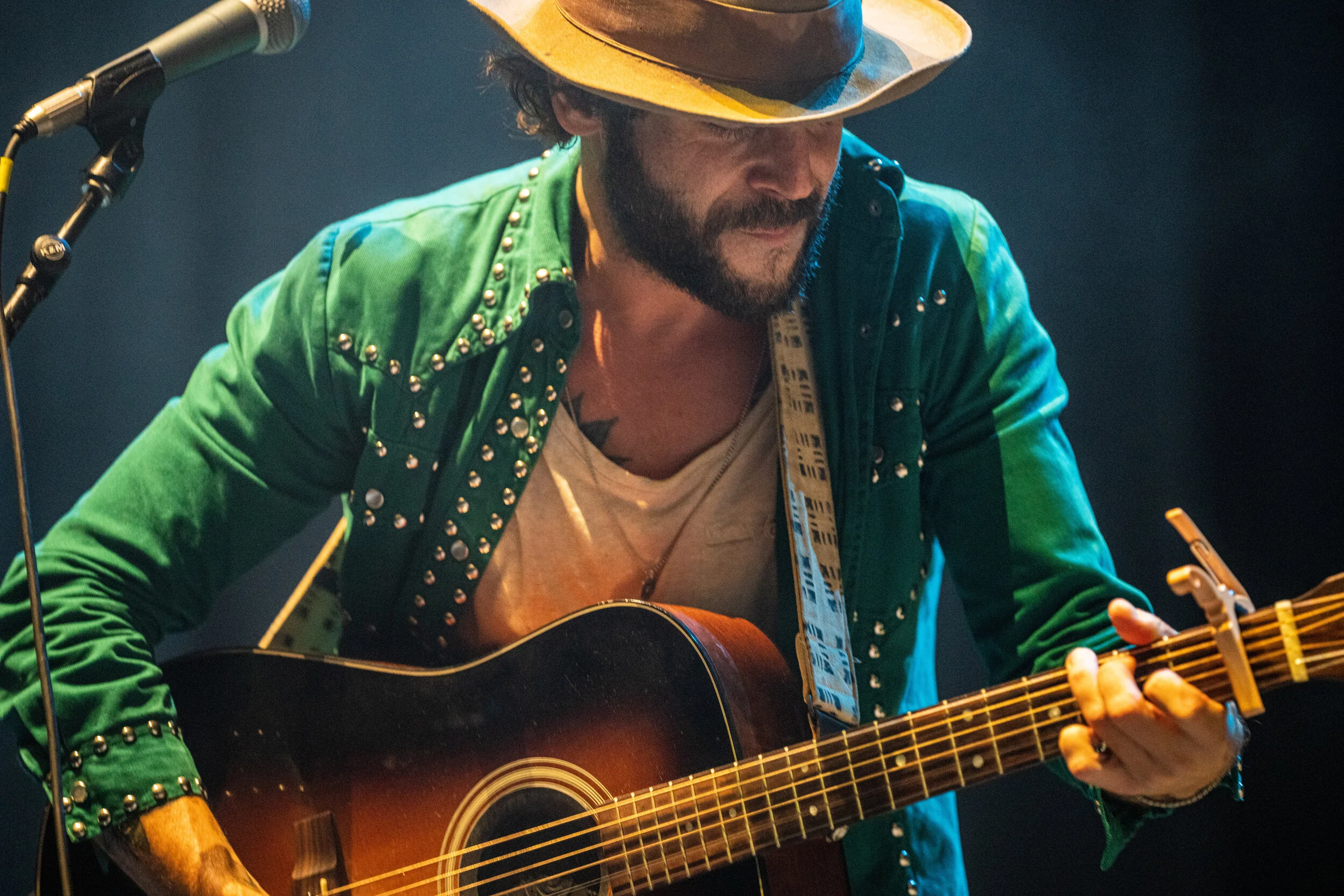 Male musician wearing a straw cowboy hat and green jacket with metal studs, playing an acoustic guitar on stage.