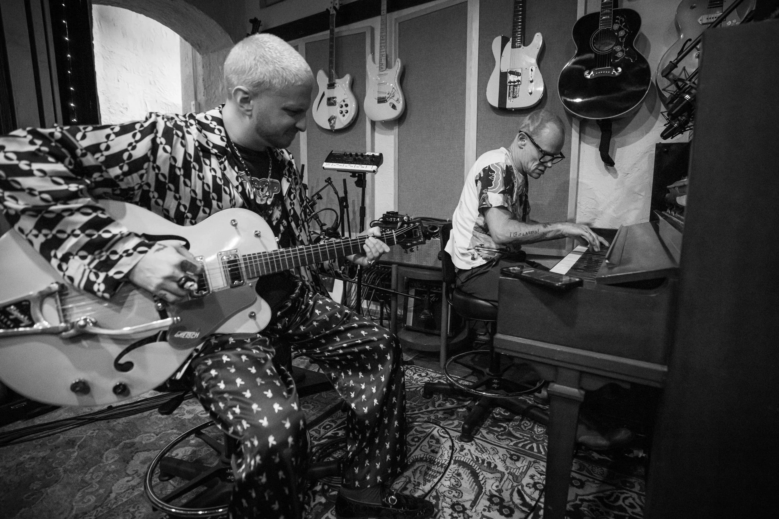 Two musicians in a music studio, one playing an electric guitar and the other playing a piano, with guitars hanging on the wall behind them.