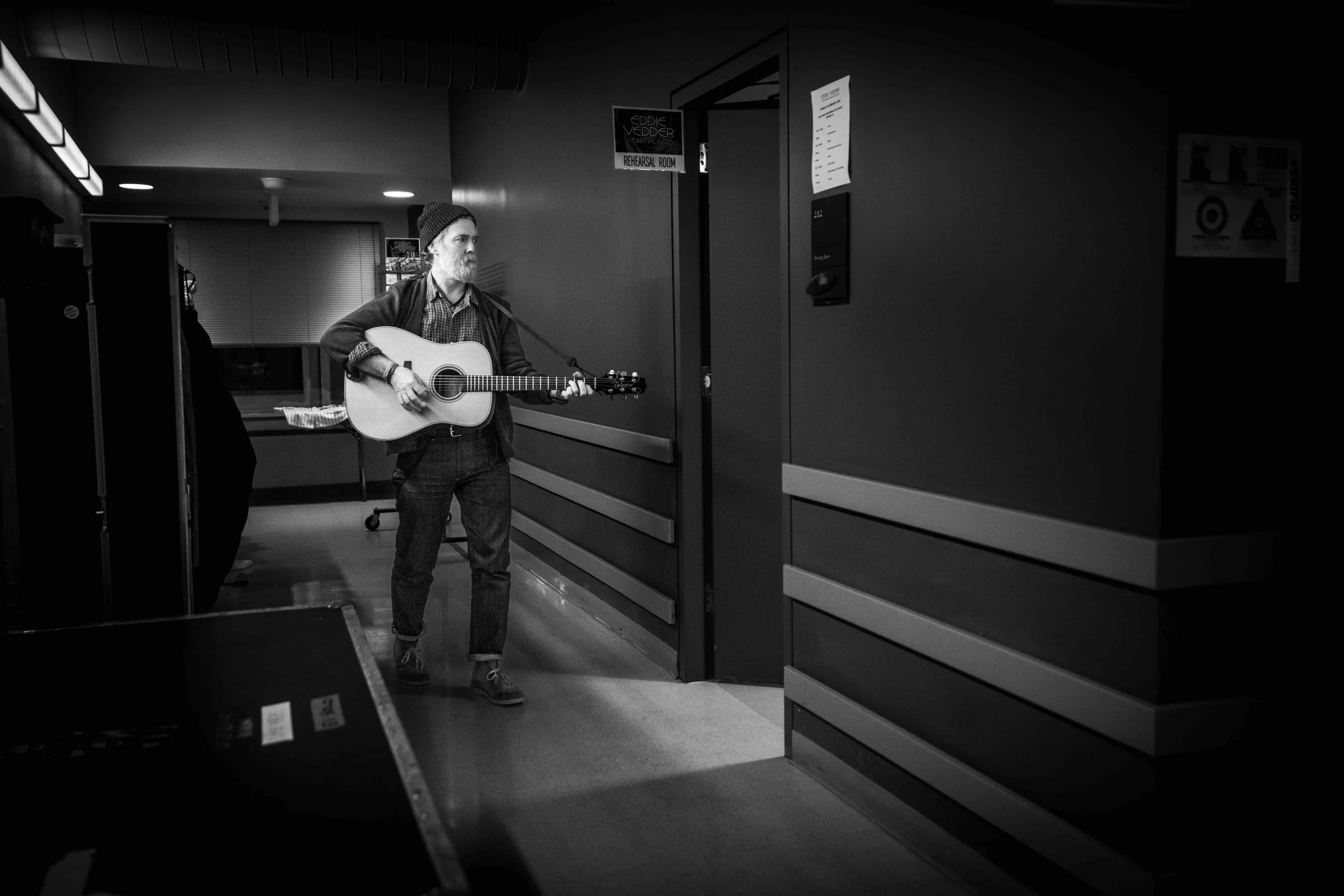 A man with a beard wearing a beanie, shirt, and jeans playing an acoustic guitar in a hallway, possibly in a rehearsal space or music venue.